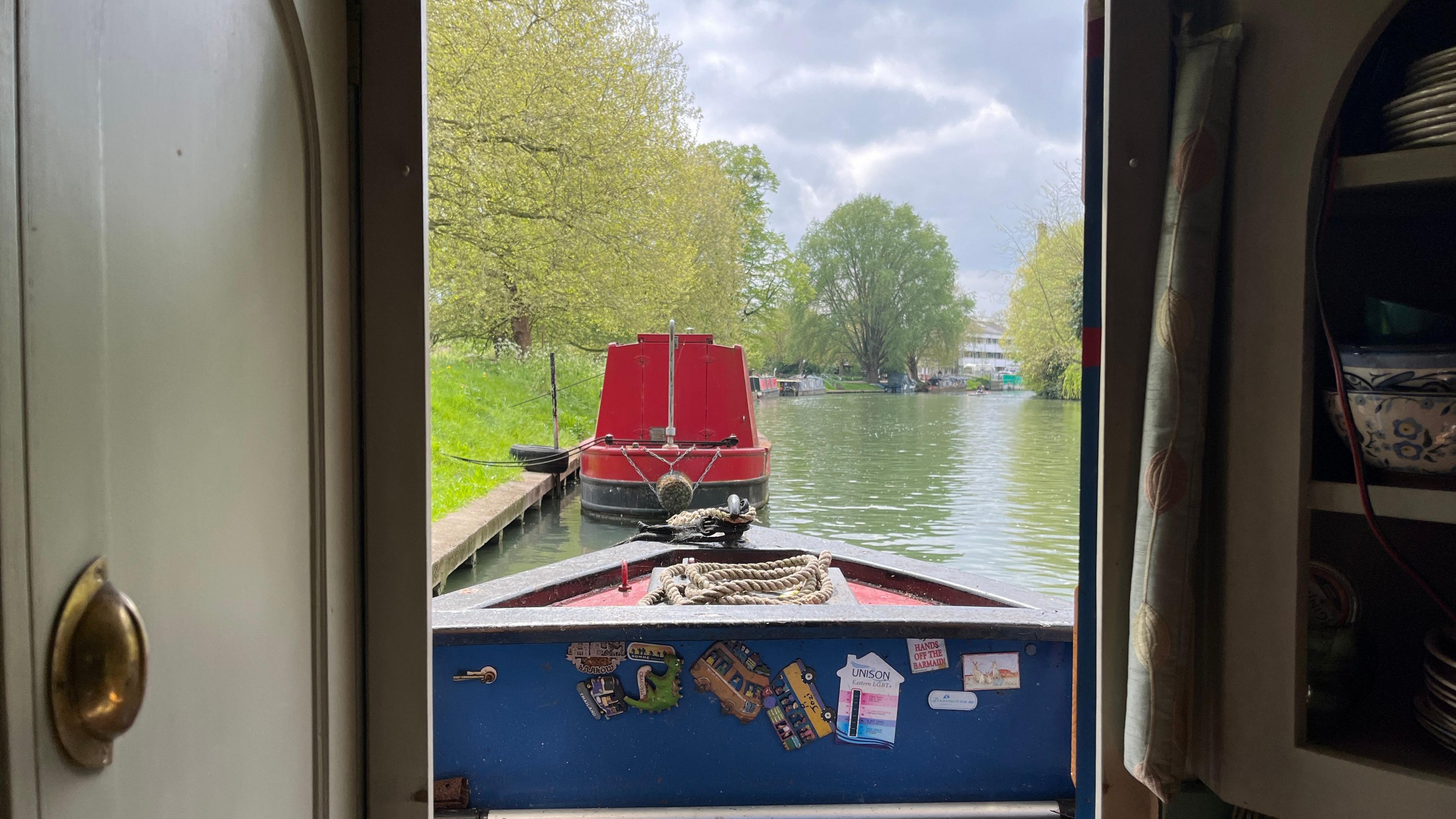 The view of a river and other moored boats of different colours from inside the thin door frame of a canal boat. There are trees and grass on the edges of the canal and the river water is a murky green colour. The sun is trying to peek through a cloudy sky. The boat directly in front of where the photo is being taken is red. It has been tied to the side of the river with a rope.
