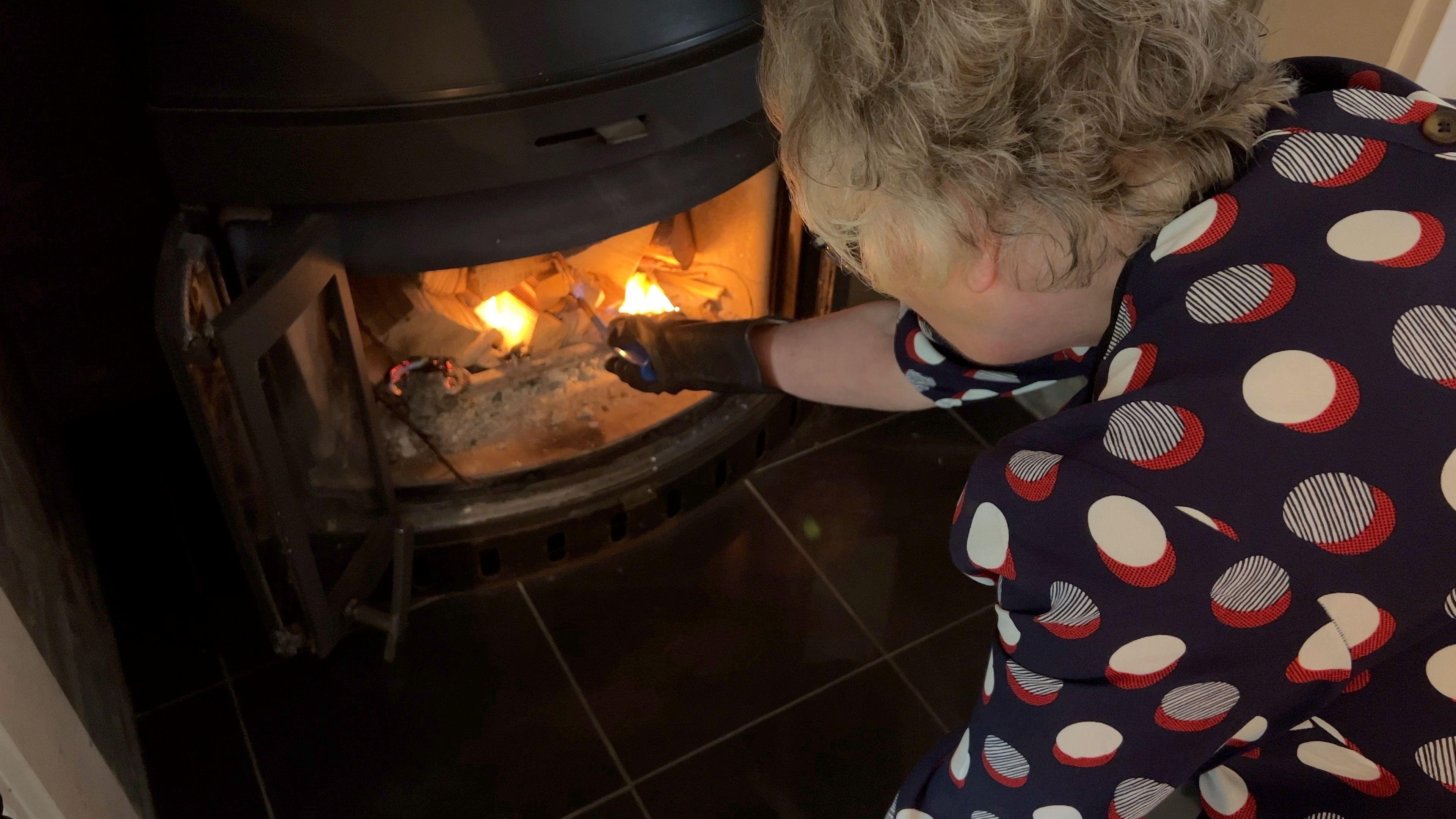 A woman with grey hair and wearing a spotty blouse stoking a fire in a log burner.