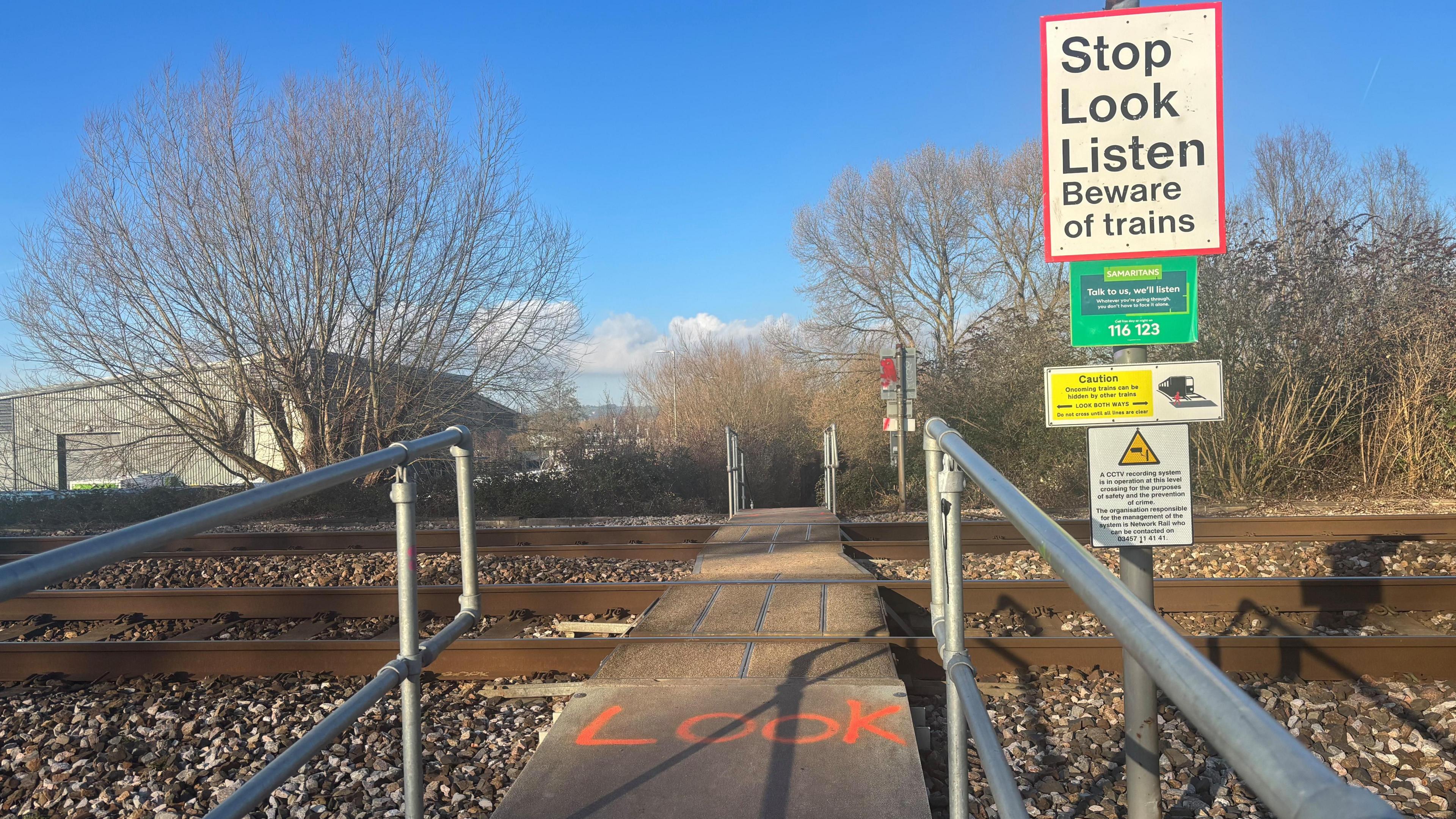 A foot crossing across a railway track. The word "Look" is spray painted on the floor and there is a sign to the right which says "Stop, Look, Listen". There is an industrial unit in the background to the left and trees to the right.
