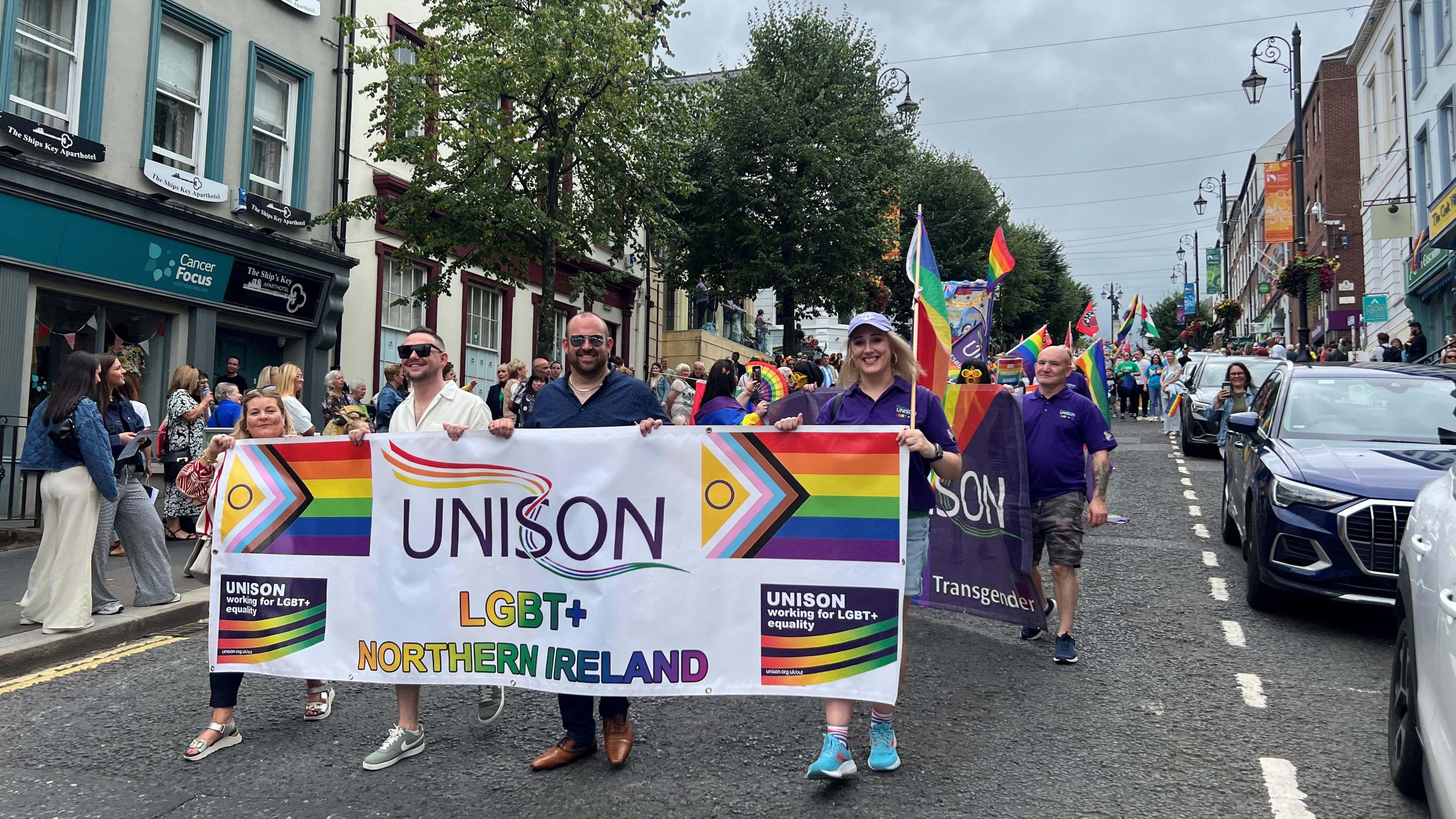 Four people are walking on a road, holding a large white banner. On the banner are the words 'UNISON LGBT+ NORTHERN IRELAND'. There are also two rainbow flags on the banner, as well as two graphics which read 'UNISON working for LGBT+ equality'. Behind the group is a crowd who are also part of the parade.