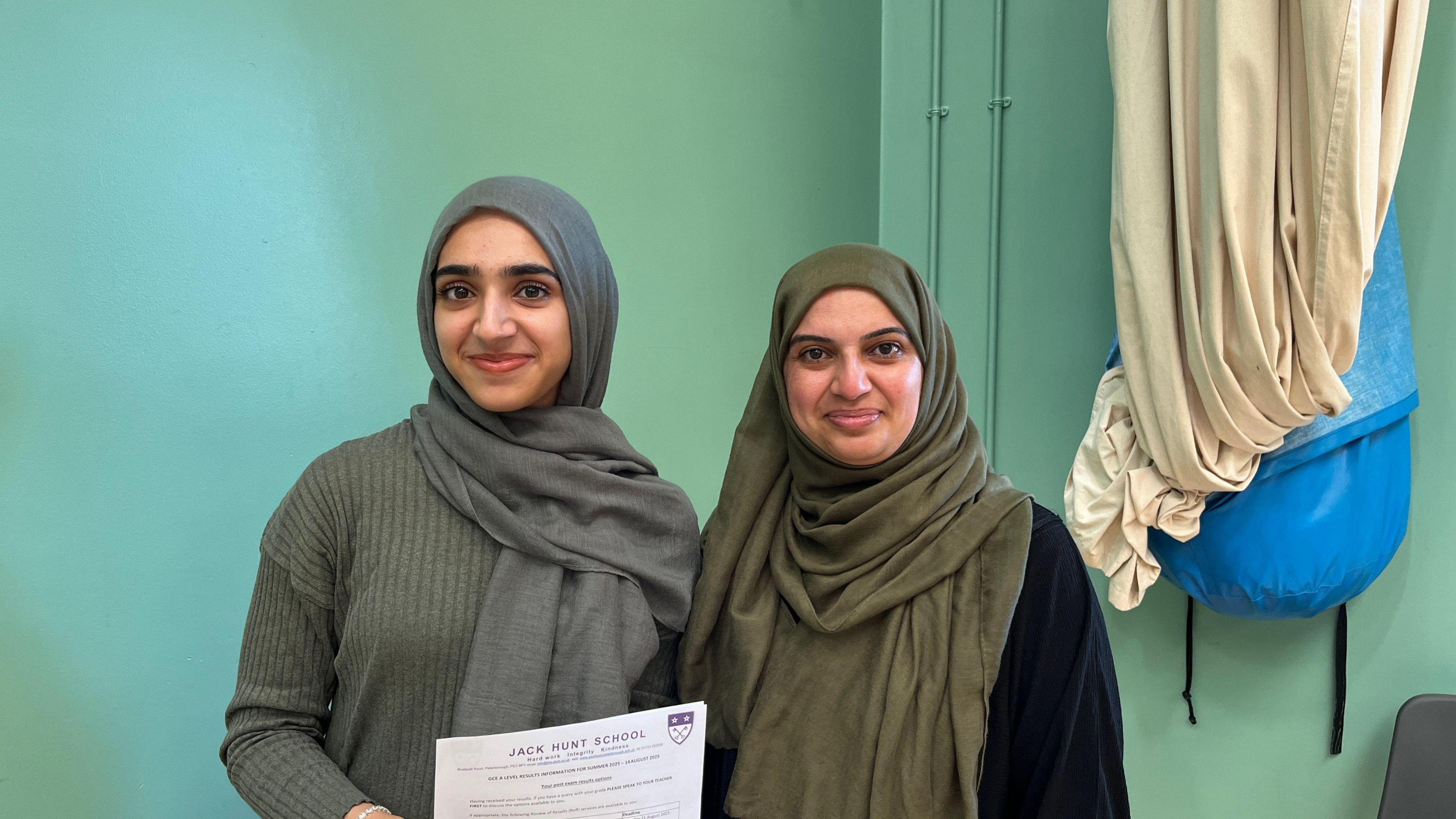 Husna, wearing a grey jumper and a grey headscarf, with her mum, in a green  headscarf, smiling for the camera.