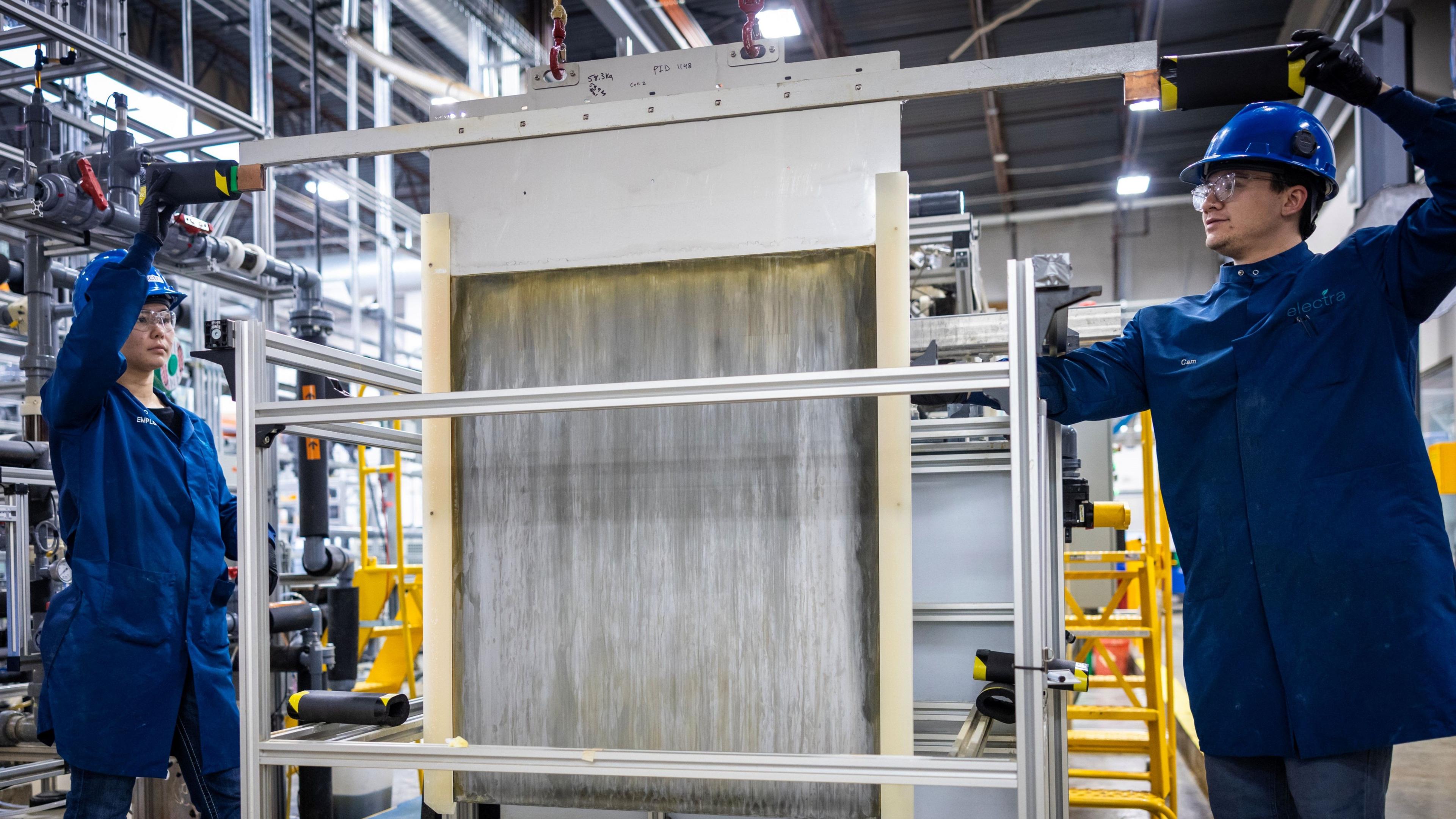 Workers in blue overalls stand each side of a frame holding a sheet of Electra's steel.