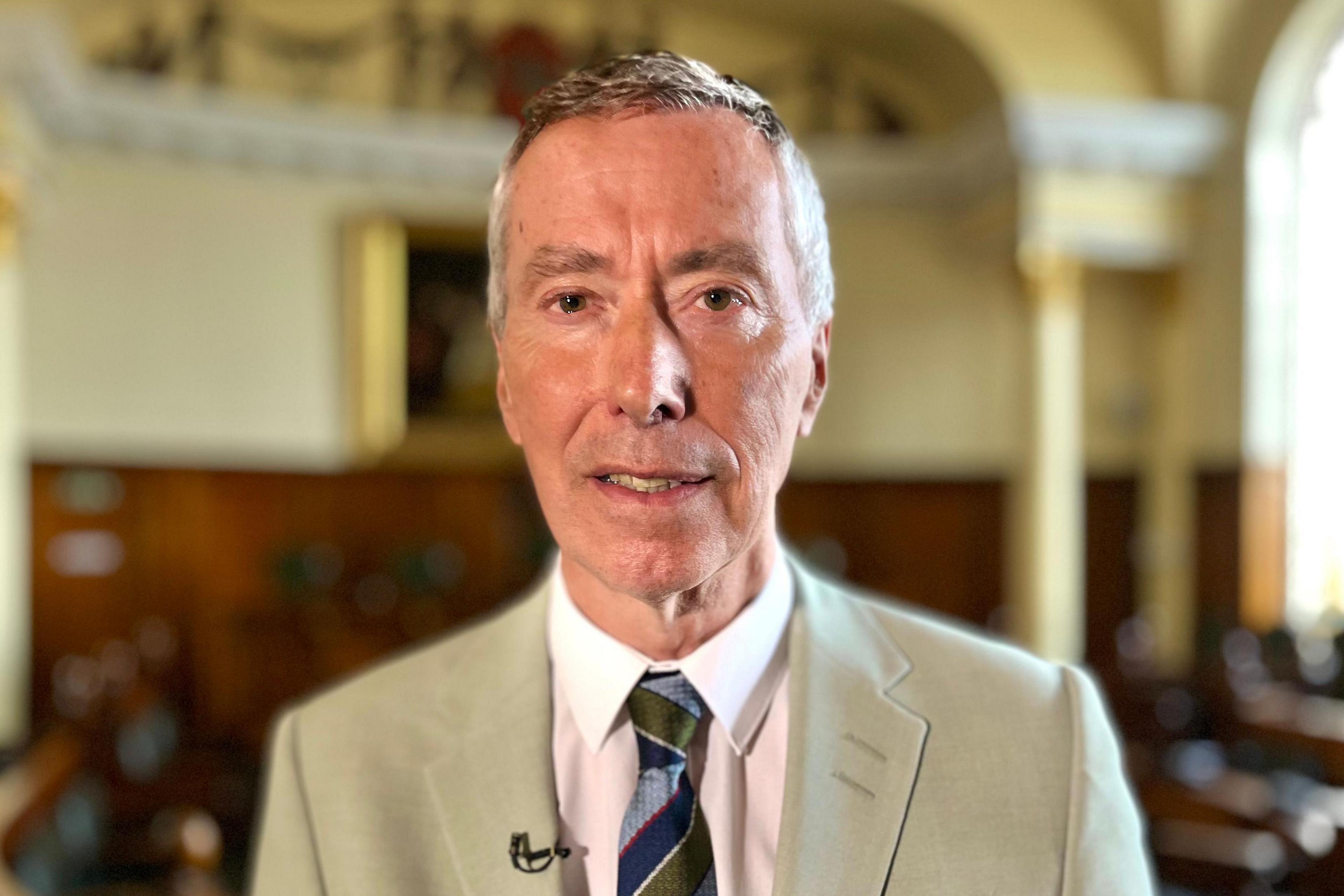 David King in a cream coloured suit and a striped tie. He has short white hair. He is standing inside a council chamber.