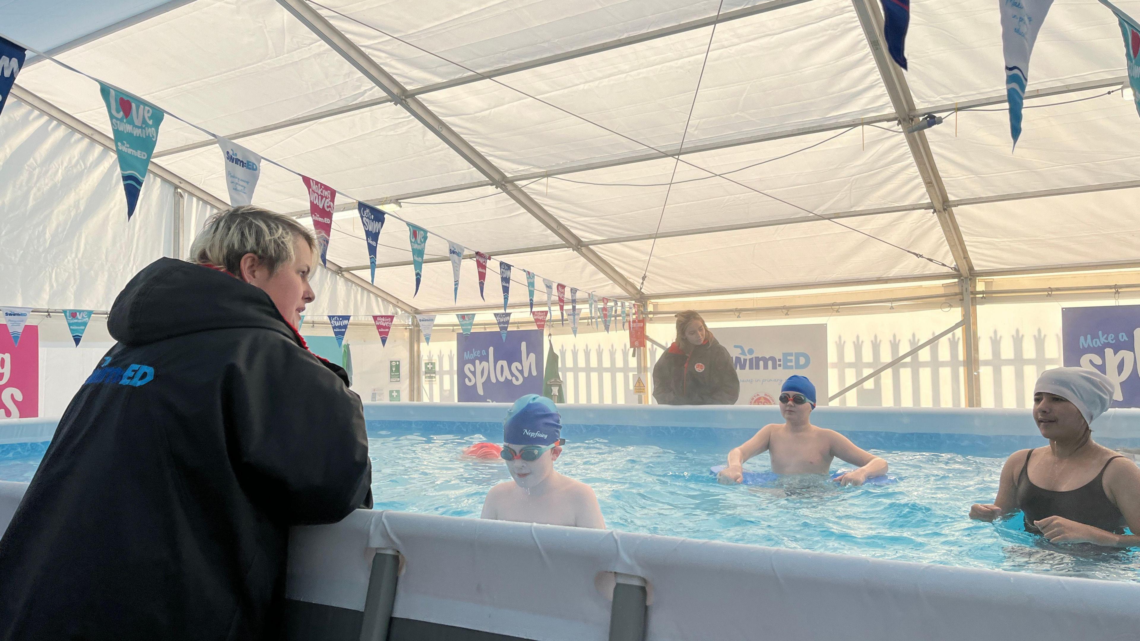 A temporary swimming pool with pupils swimming in it. A lady in a black hoodie is leaning on the side of the pool coaching the students.