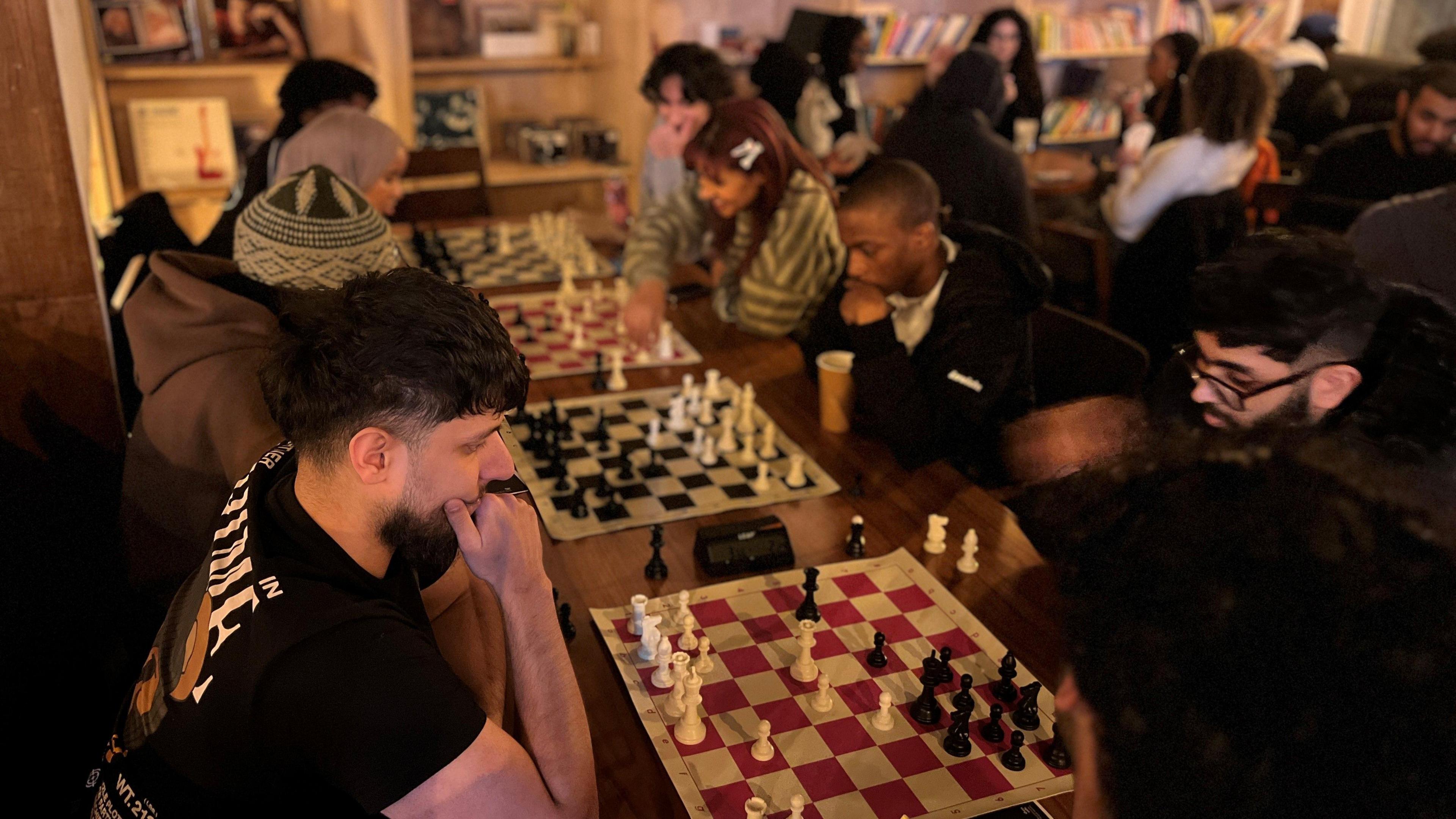 A group of young men and woman are sitting in two lines at a table, at opposing sides. Chess boards are in the middle and they are all staring down at them and looking thoughtful.