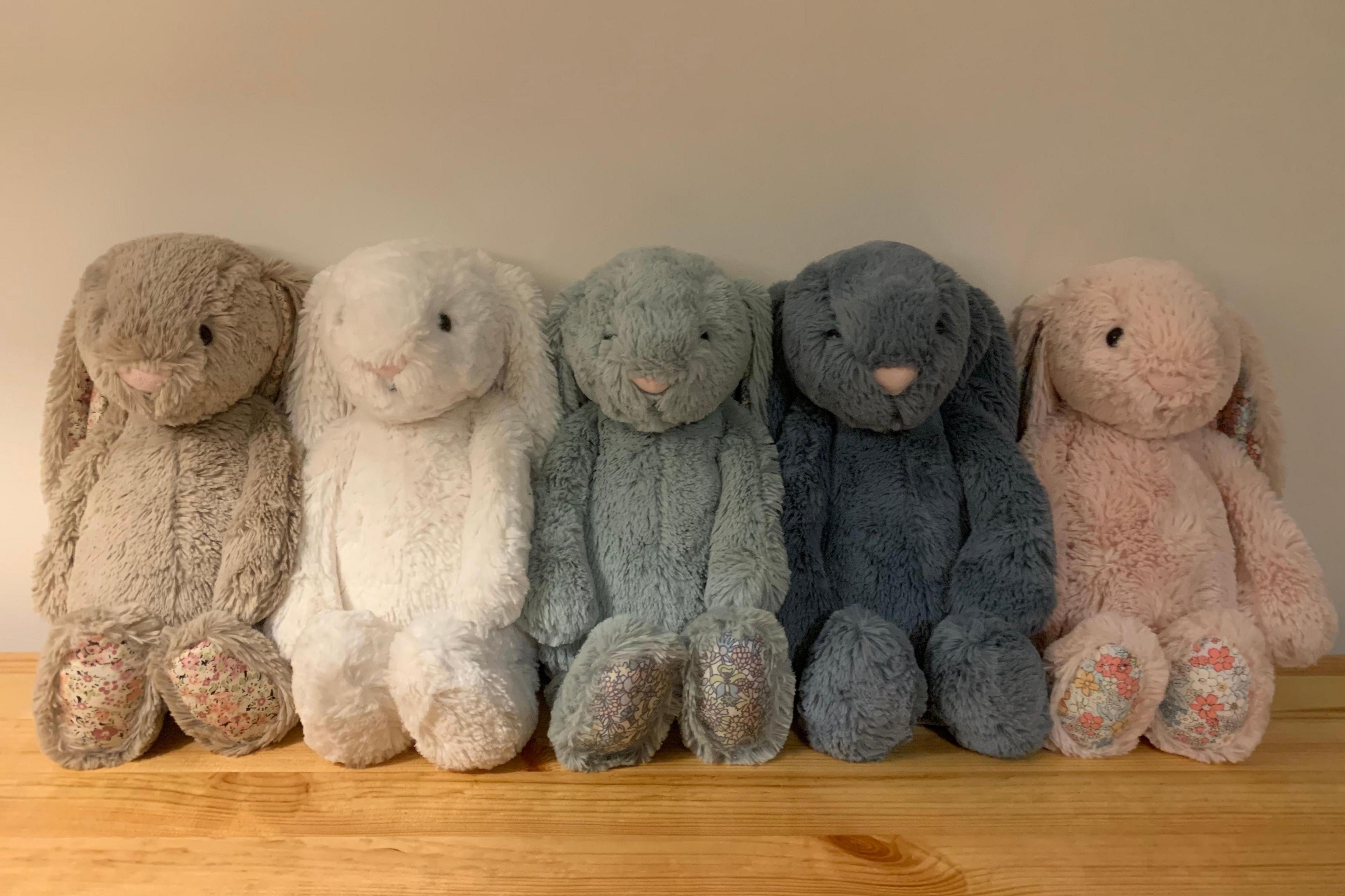 Five cuddly rabbit type teddy bears known as Jellycat bunnies sit in a row, side by side on a brown mantelpiece. One is a fawn colour, the others are white, light blue, dark blue and pink.


