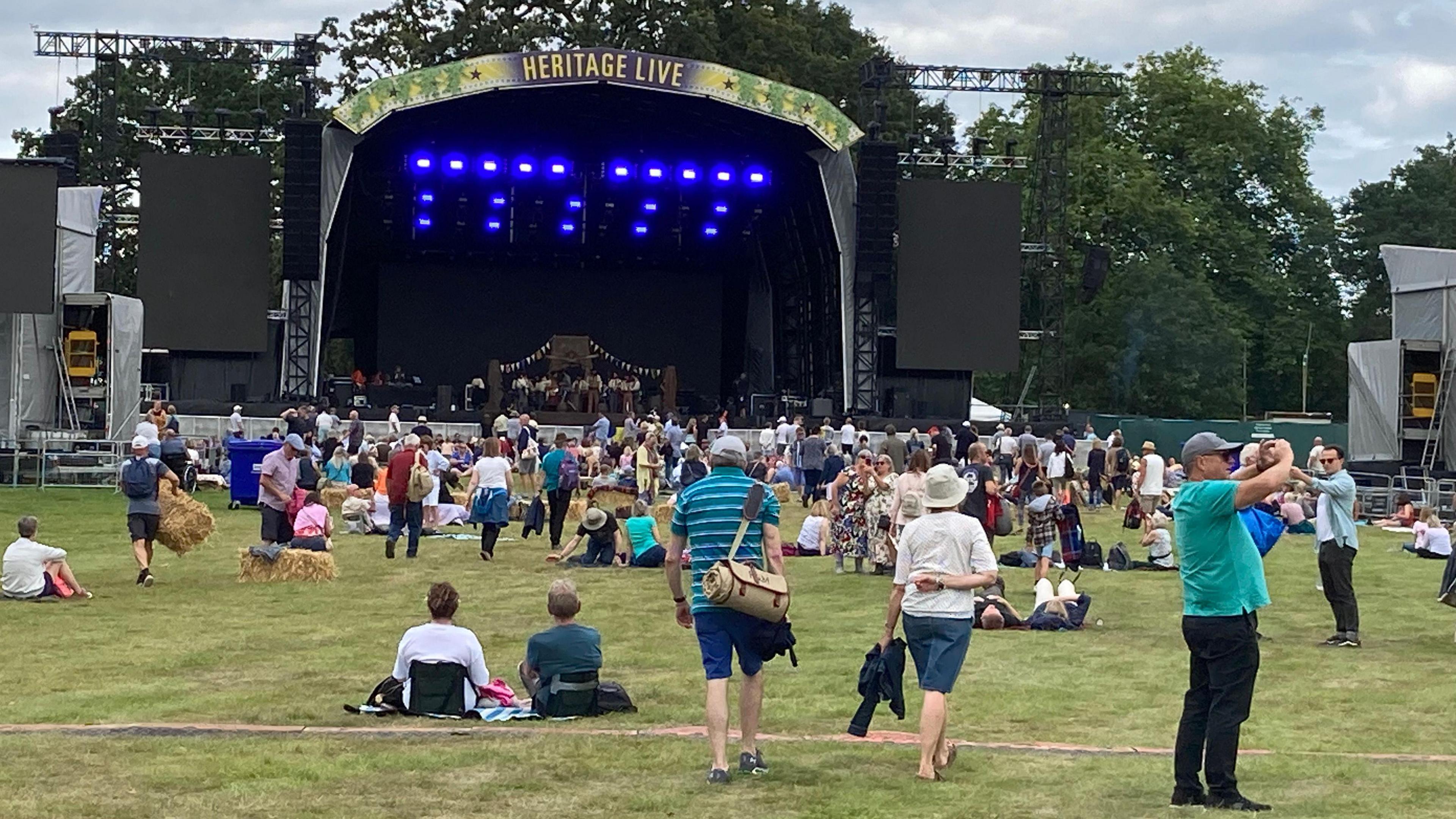 People in front of an outdoor stage sitting on grass or walking. There is a "Heritage Live" logo above the stage.  There are 20 blue lights in a pattern on the backdrop of the stage. There are large black speakers on either side of the stage.