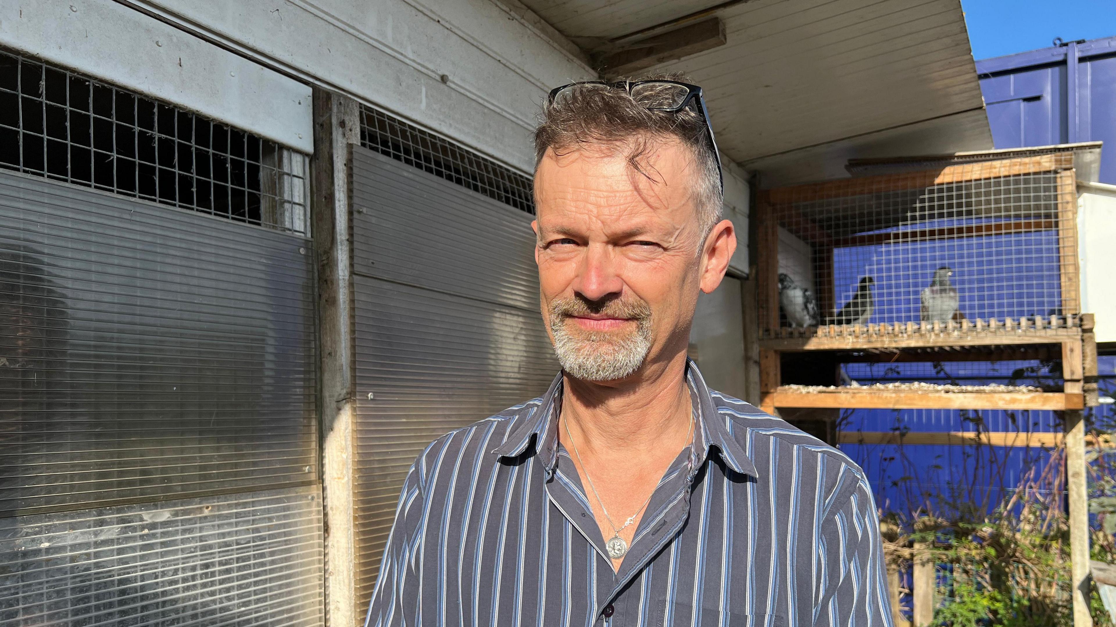 Stuart is staring at the camera. He has grey hair and is wearing a blue striped shirt and behind him are a number of pigeon lofts