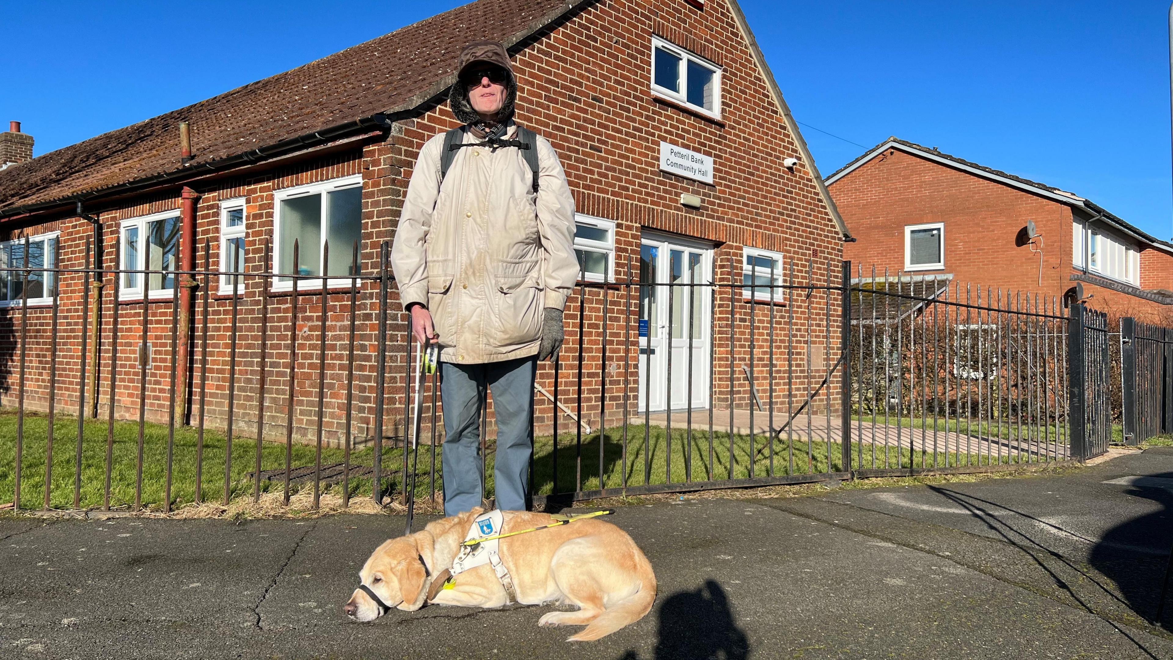 John Atkinson is standing outside his local polling station in Carlisle. He is wearing a brown hat, grey coat and dark glasses. His guide dog Grace is at his feet and he's holding her lead.