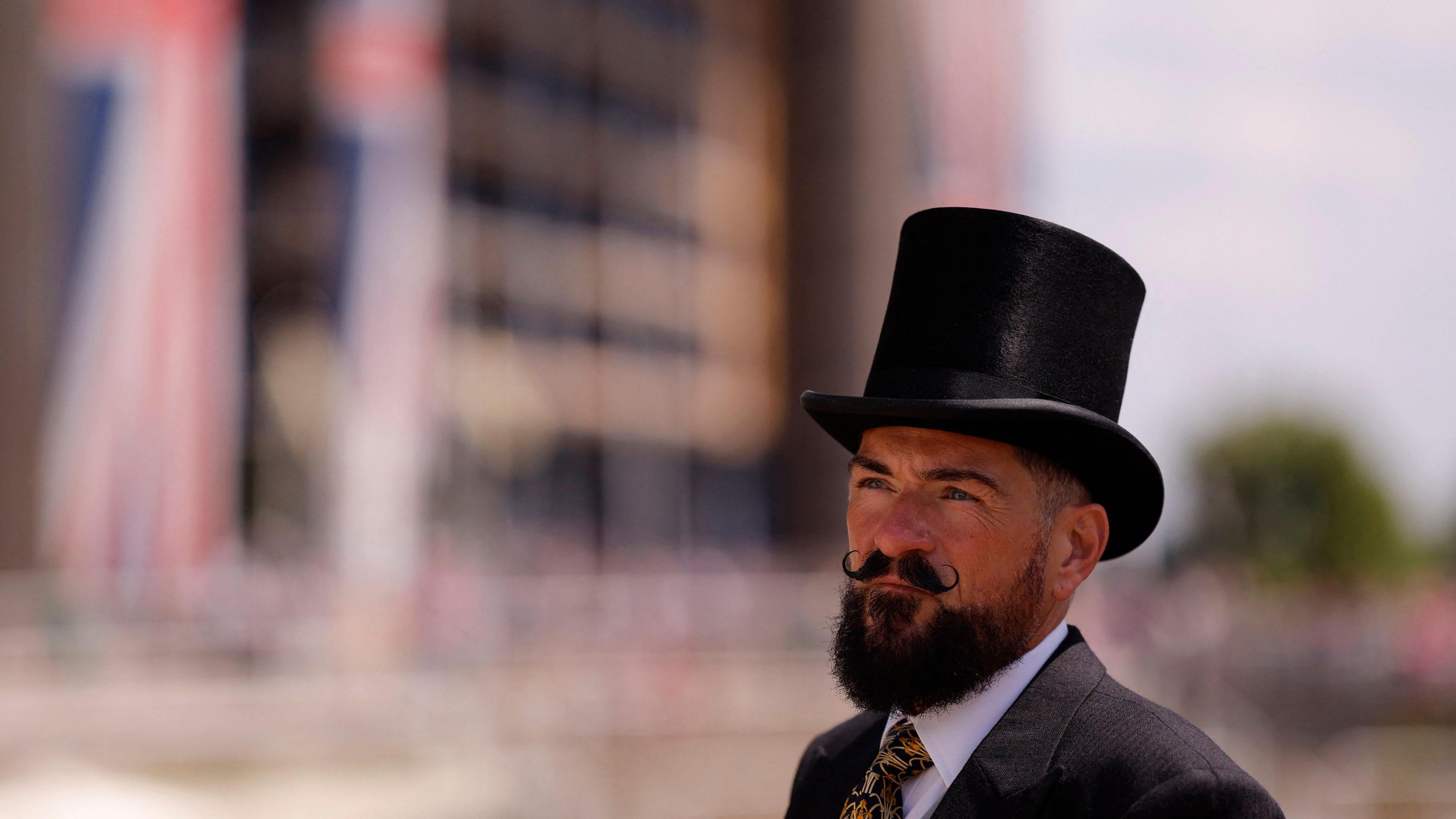 A man looks away from the camera, wearing a black top hat, white shirt, tie and blazer, with a full beard and moustache