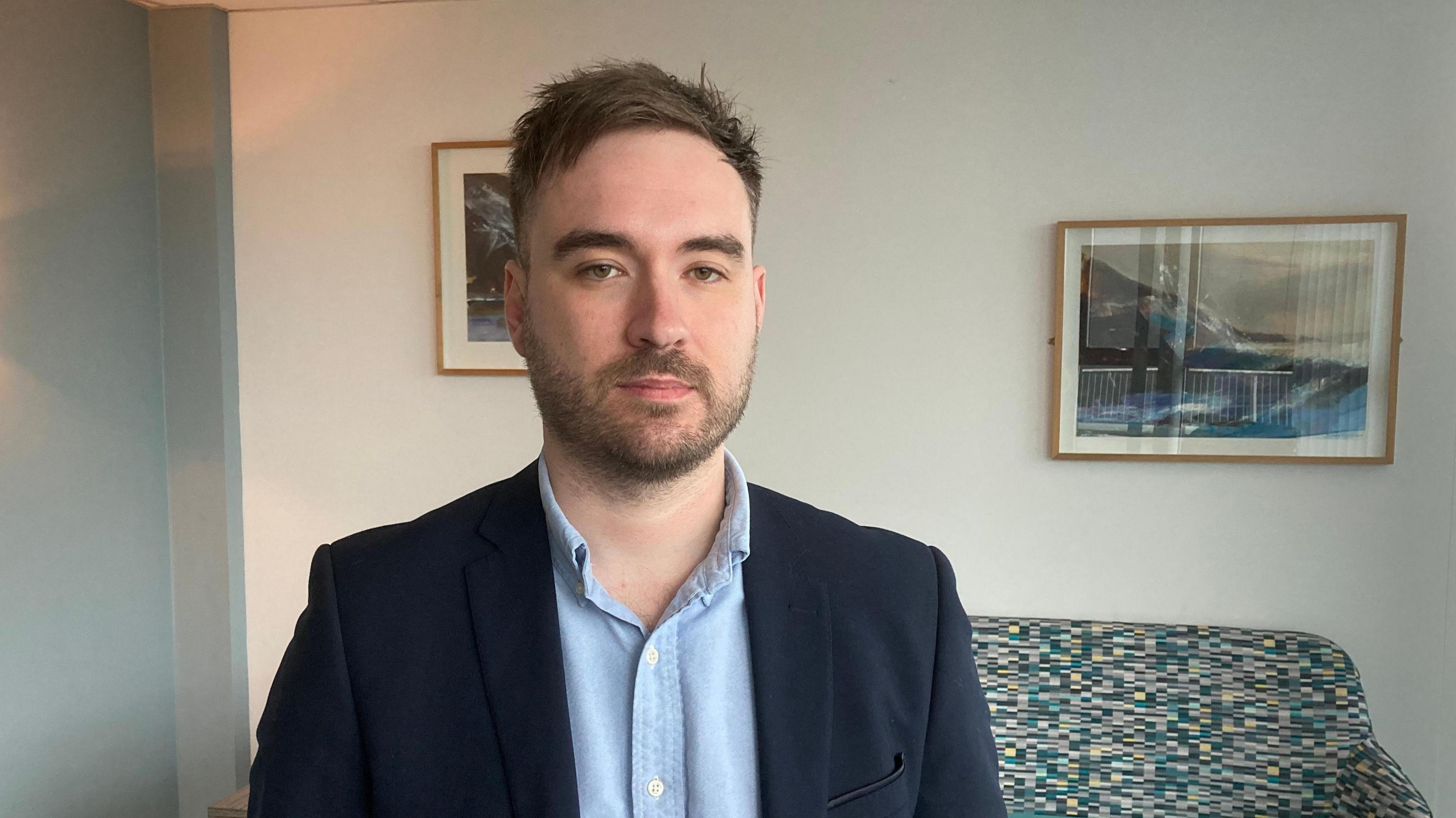 Dr Jamie Torrance - he is wearing a navy blue suit jacked and a light blue shirt with the top button undone. A multi-coloured sofa is visible behind him and there are framed photos of mountain landscapes on the wall 