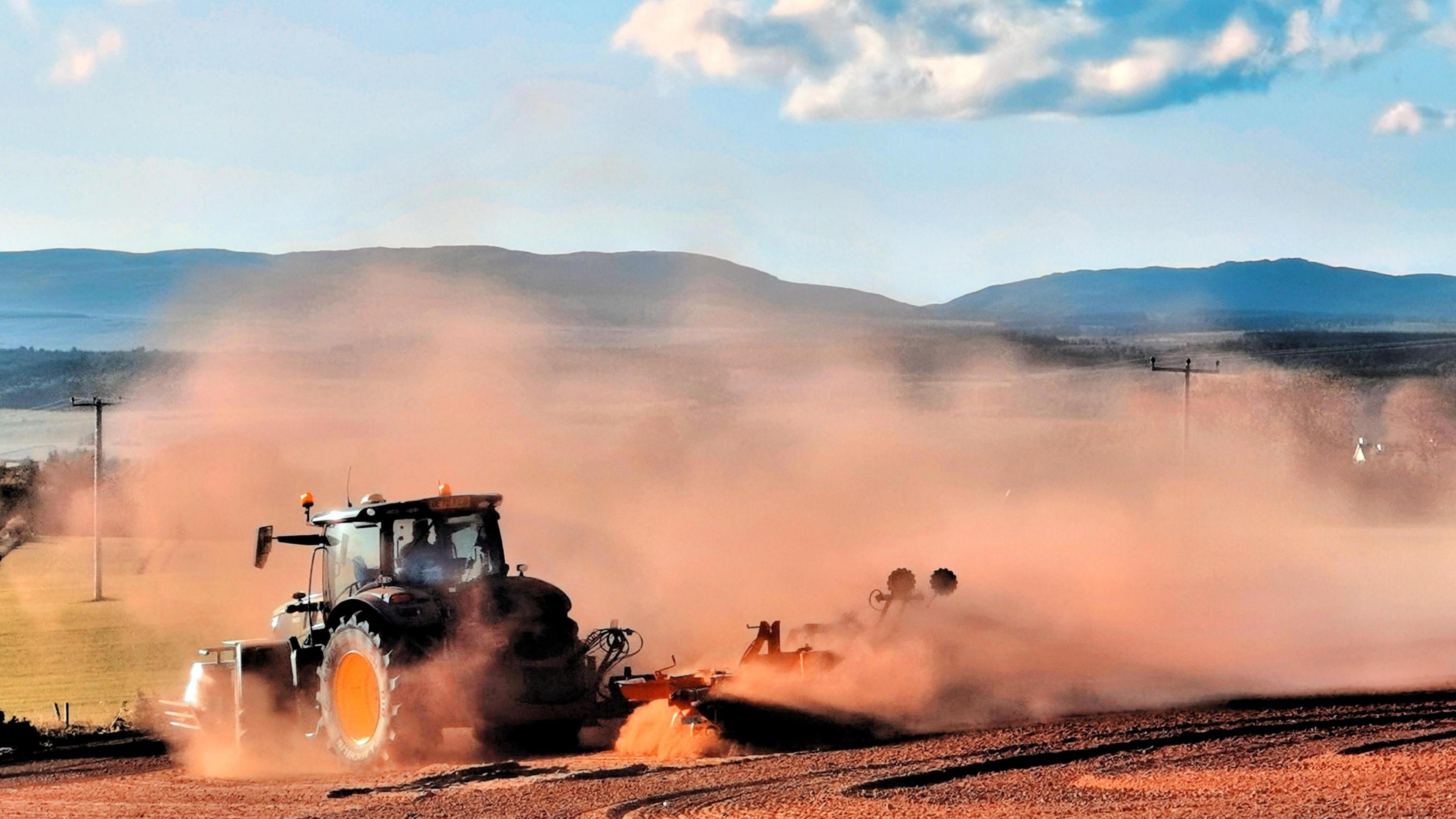 A tractor pulling equipment throws up clouds of dust as it drives through a field, with mountains and blue sky in the background