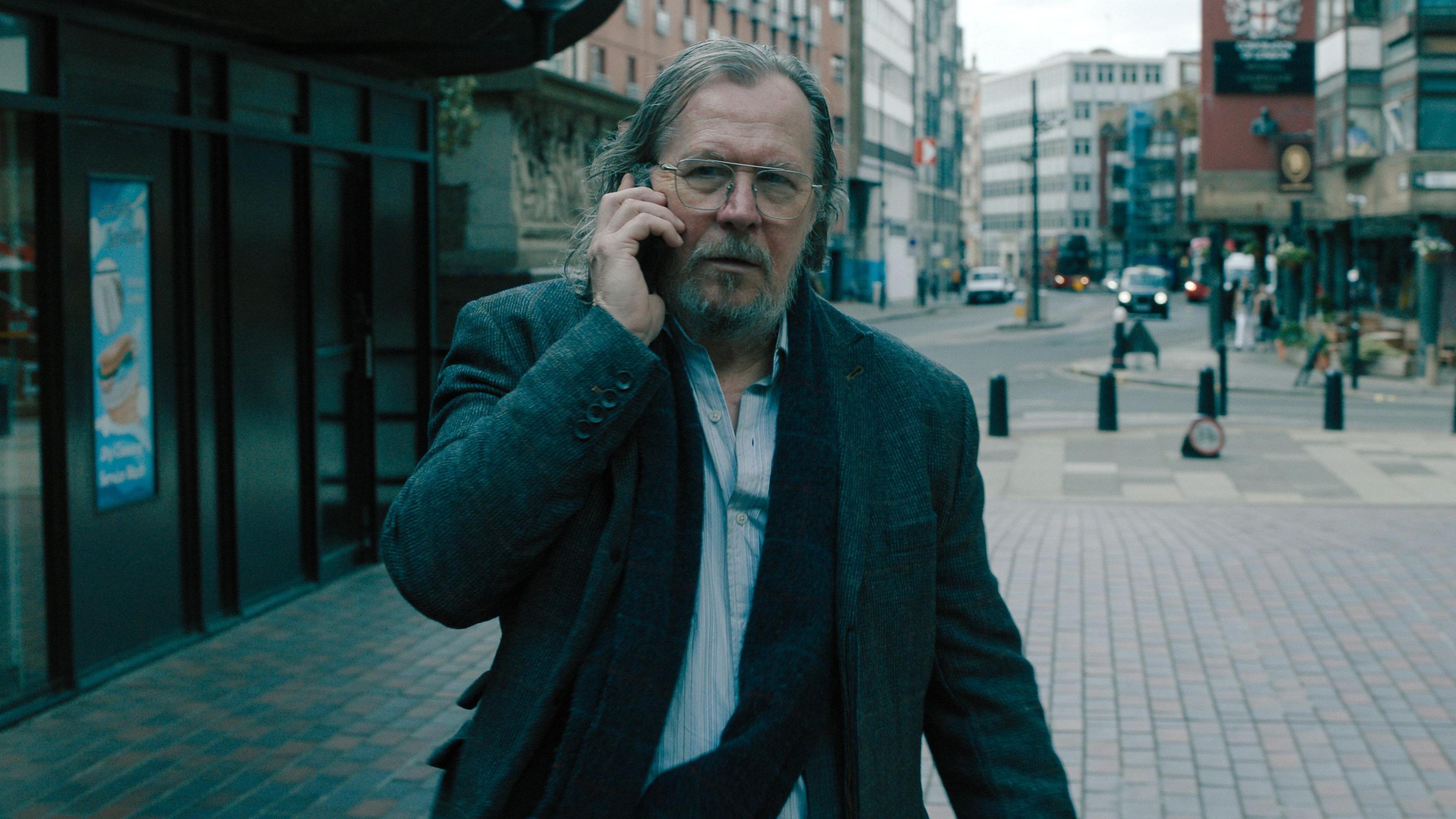 Gary Oldman has long hair and grown out facial hair. He is on his phone walking through a quiet area of a city with buildings surrounding him.