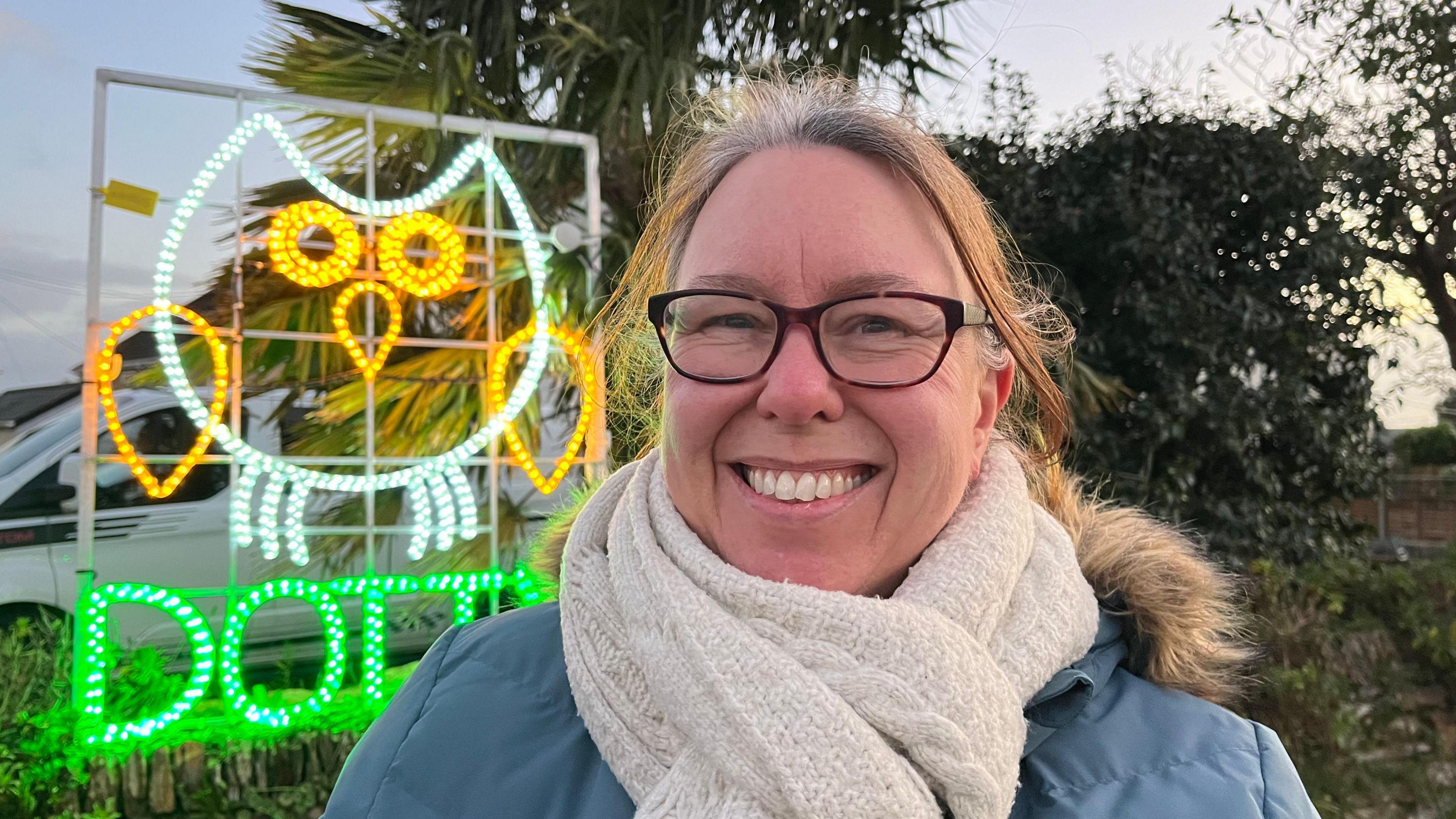 A yellow and green Christmas light display of an owl with the word Dotty below, with a woman smiling in front of it