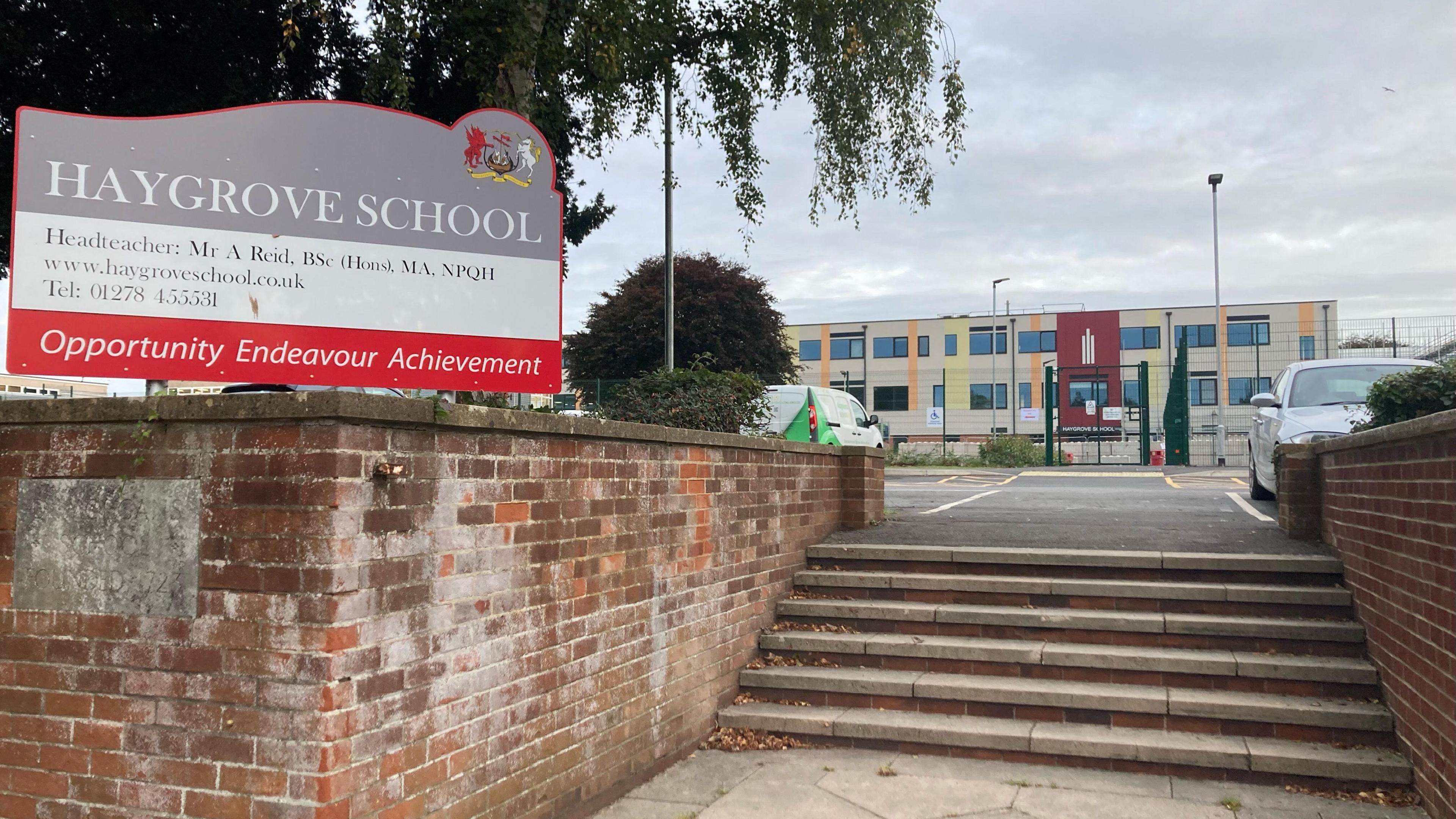 Haygrove School in Bridgwater.  A grey sign with the school's name can be seen to the left of the frame. It also features the headteacher's name, the school website and telephone. The bottom of the sign has a red stripe reading "opportunity, endeavour, achievement". The school building can be seen in the distance, behind a gate. A car and a van can be seen parked in front of the gate. 