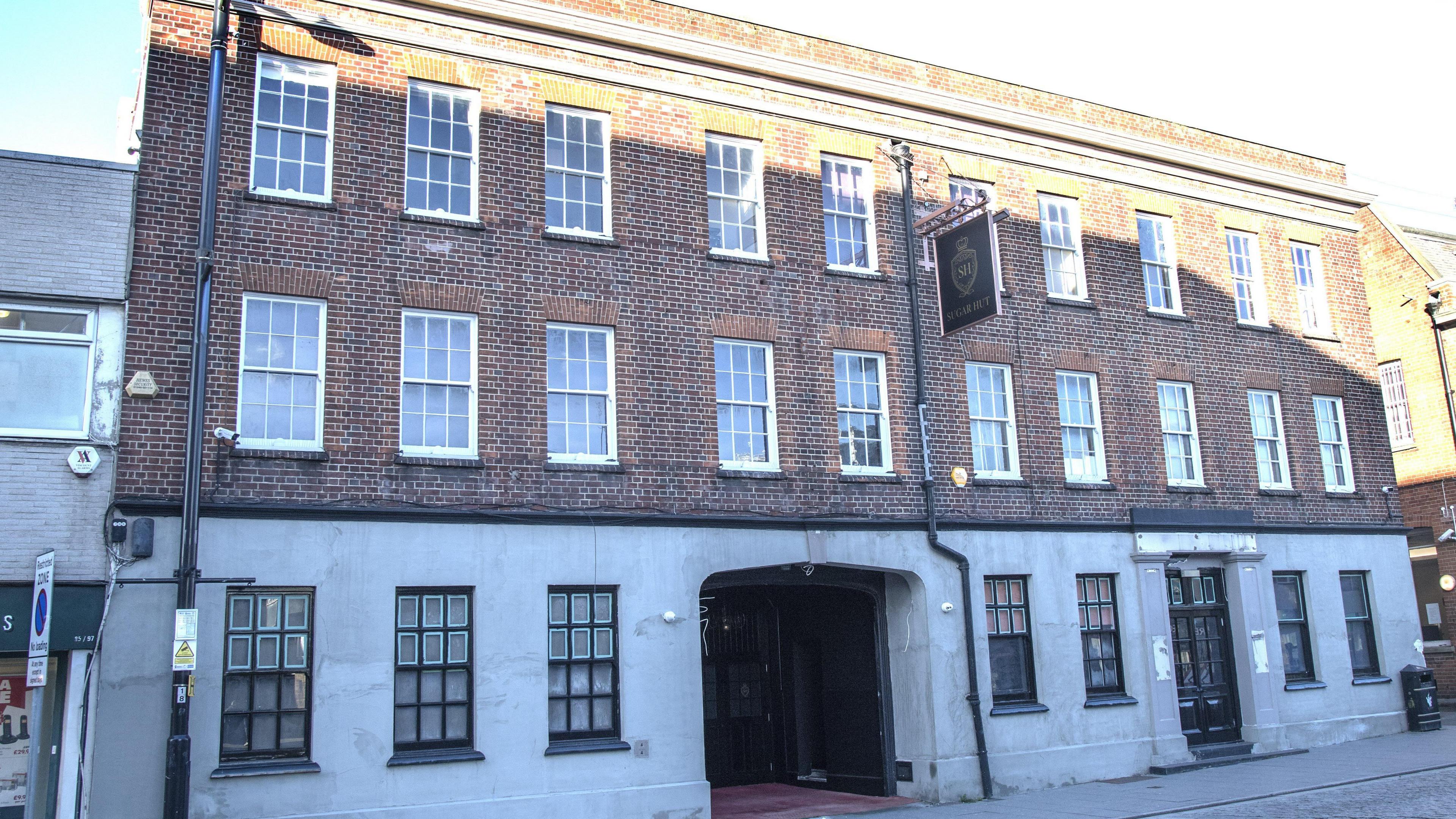 A three story building. The first floor is grey rendering and the top two are red brick. The doorway has a tall and wide arch with double wooden doors inside the arched entry. The Sugar Hut sign is on the front of the building.