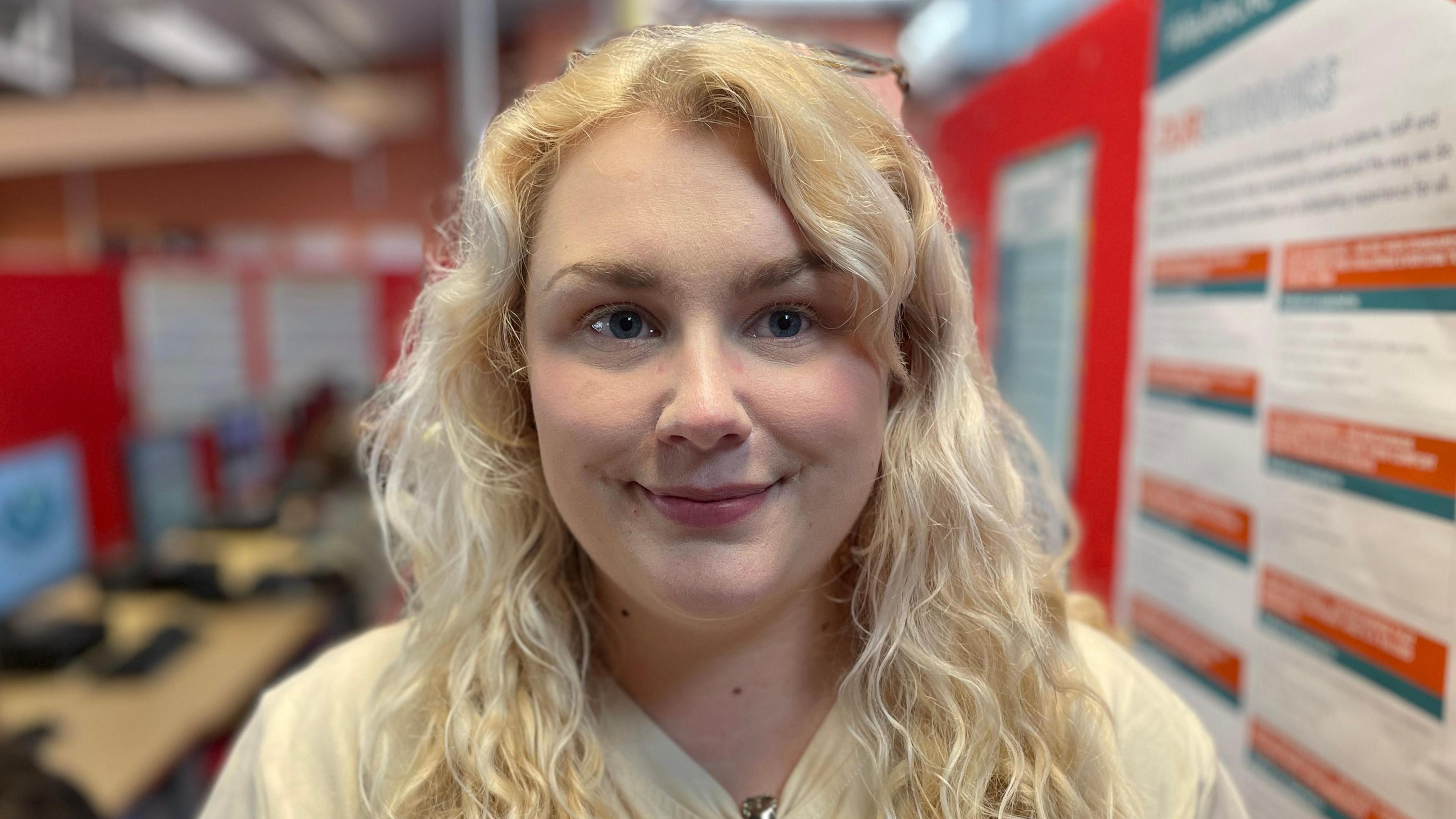 Charlotte wears her blonde, curly hair down with a white top. She is standing in a room with computer screens and red boards.