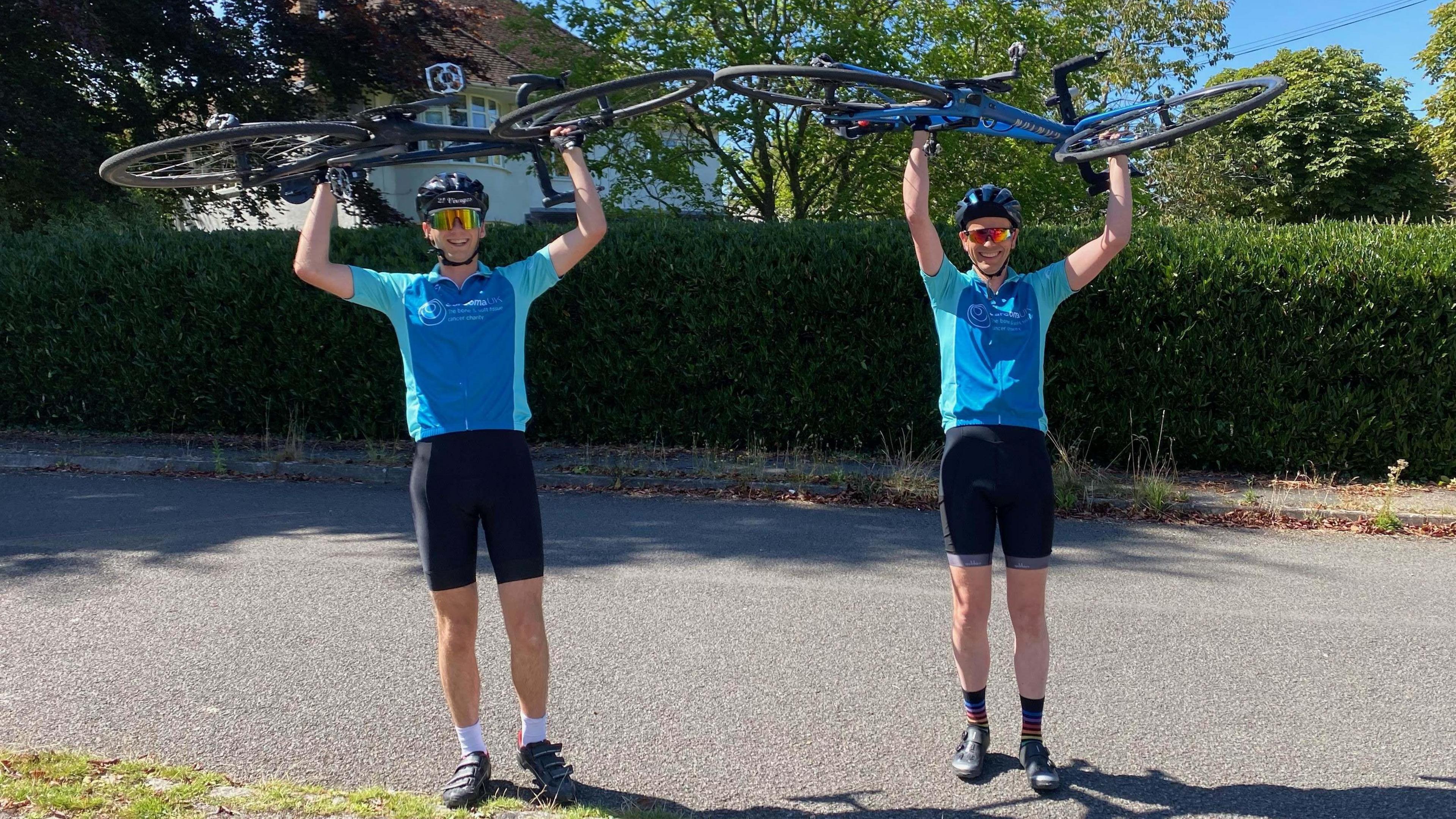 Jasper and Dominic (left and right) are both holding their bikes above their heads, wearing cycling shorts and lycra turquoise tops with helmets and sunglasses.