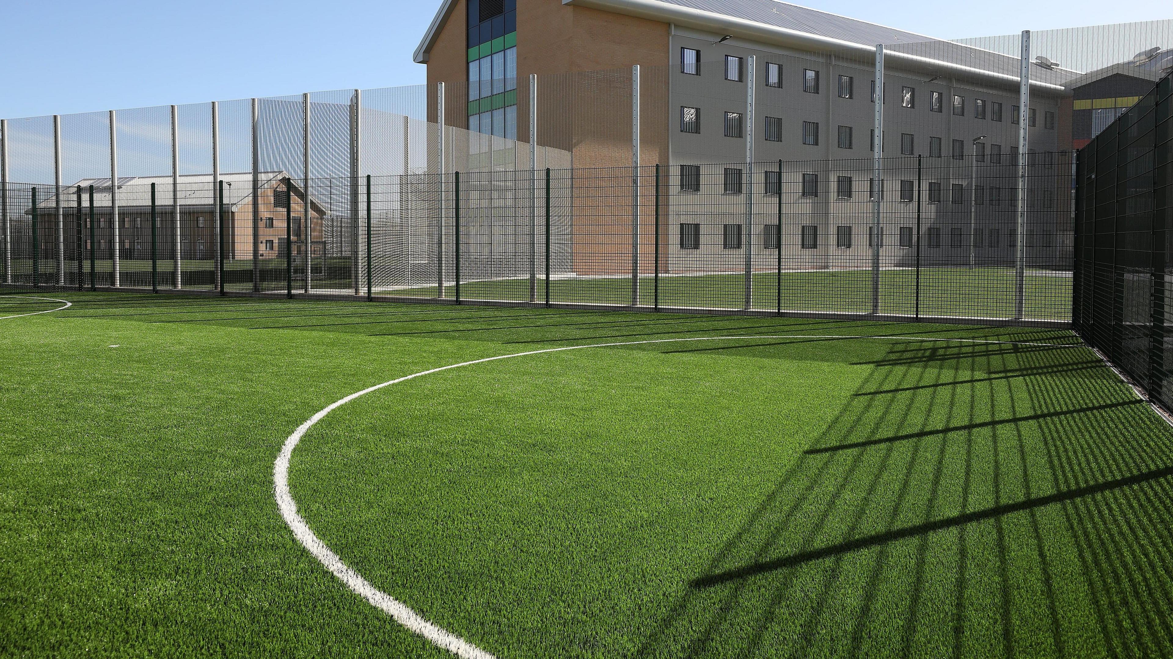 A football pitch at HMP Berwyn. An Astroturf surrounded by a white metal fence is overlooked by a prison building behind it.