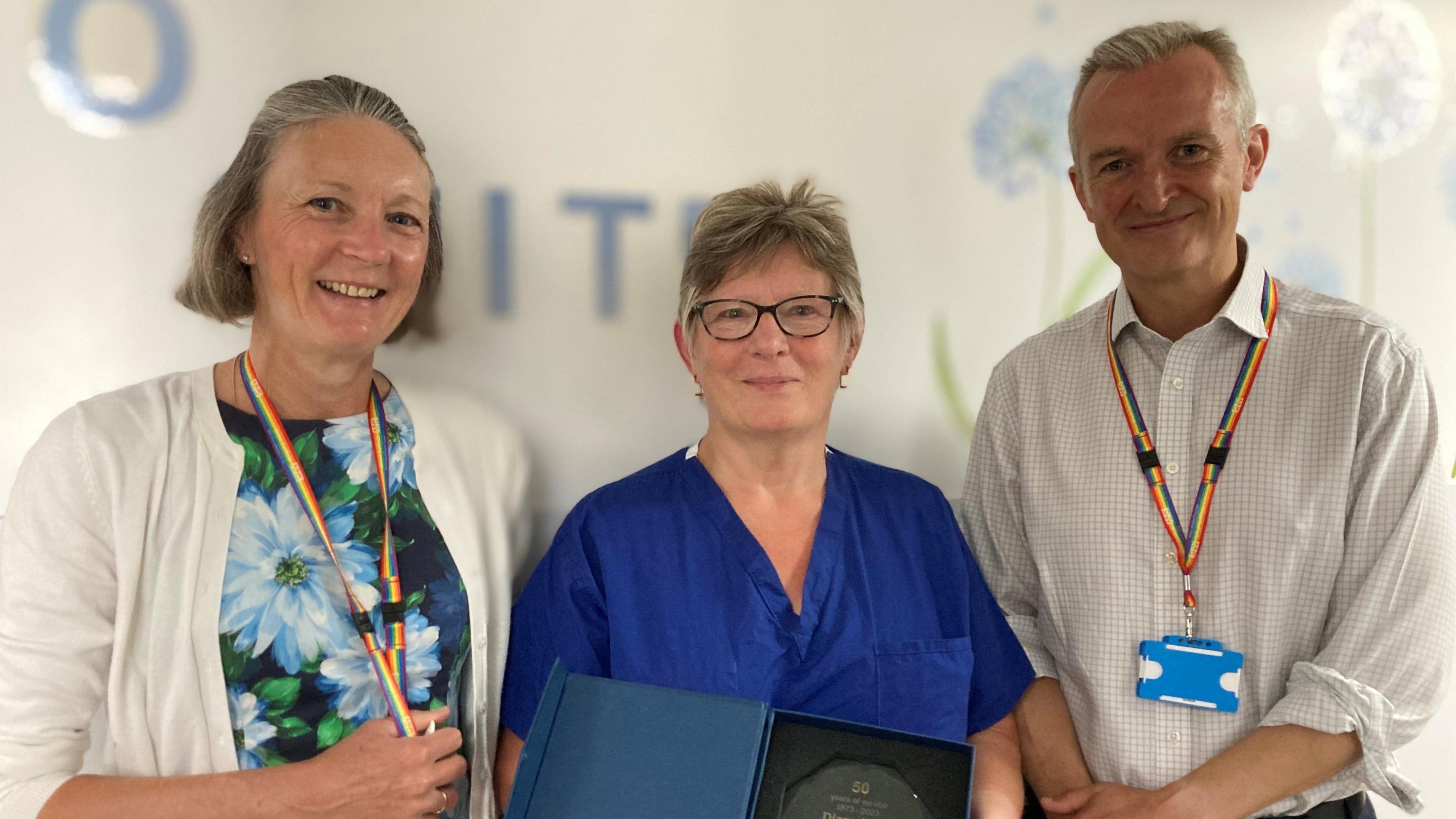 Diane Hele stands in the middle of two colleagues, she is wearing blue scrubs and holding up a glass trophy. Her colleagues beside her (a man and a woman) are smiling at the camera and both wearing lanyards. 