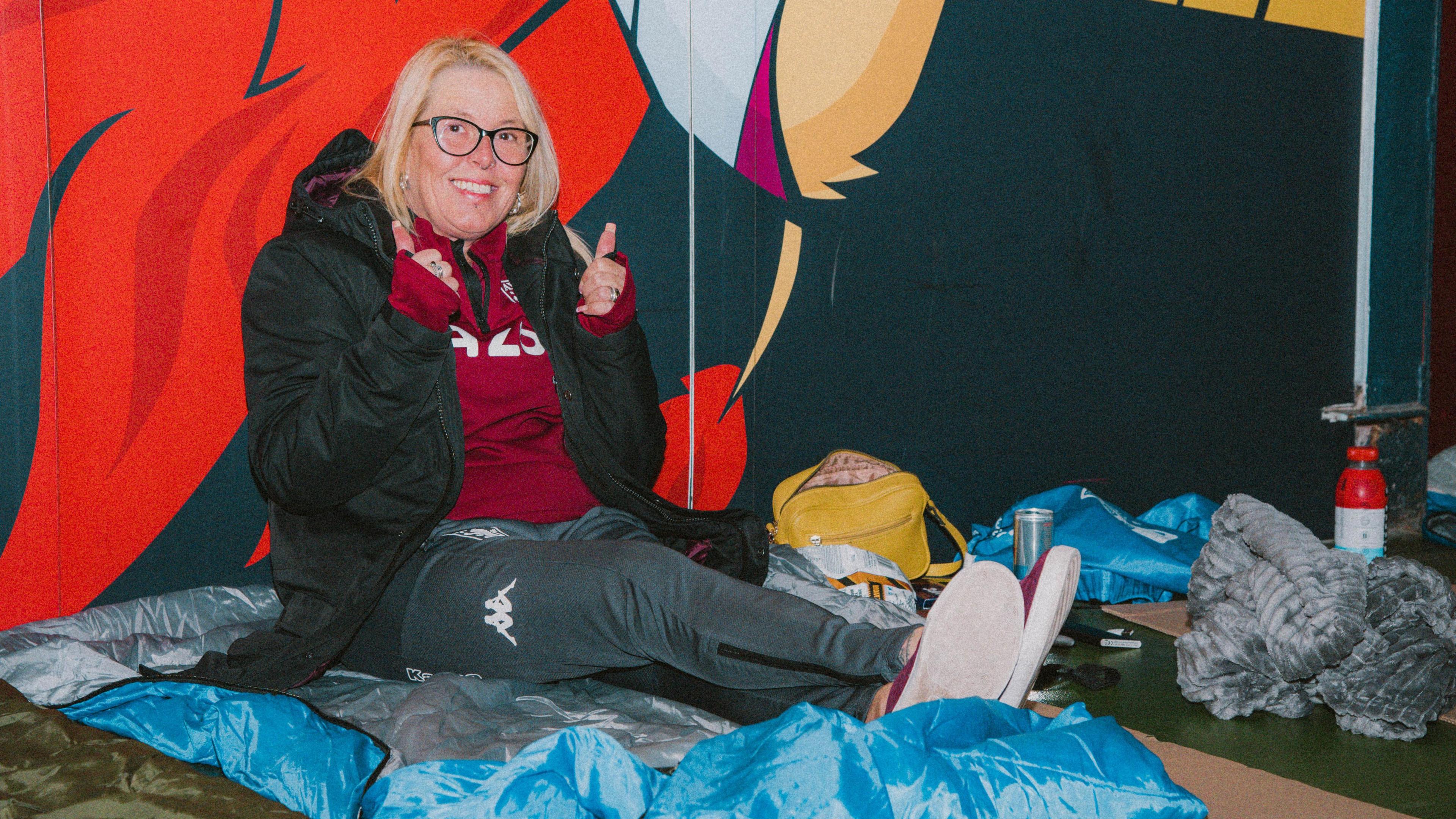 A woman with blonde hair sits on a sleeping bag in the stadium at a previous sleep out and make a thumbs up sign at the camera. She has got bags and flasks and a blanket next to her.