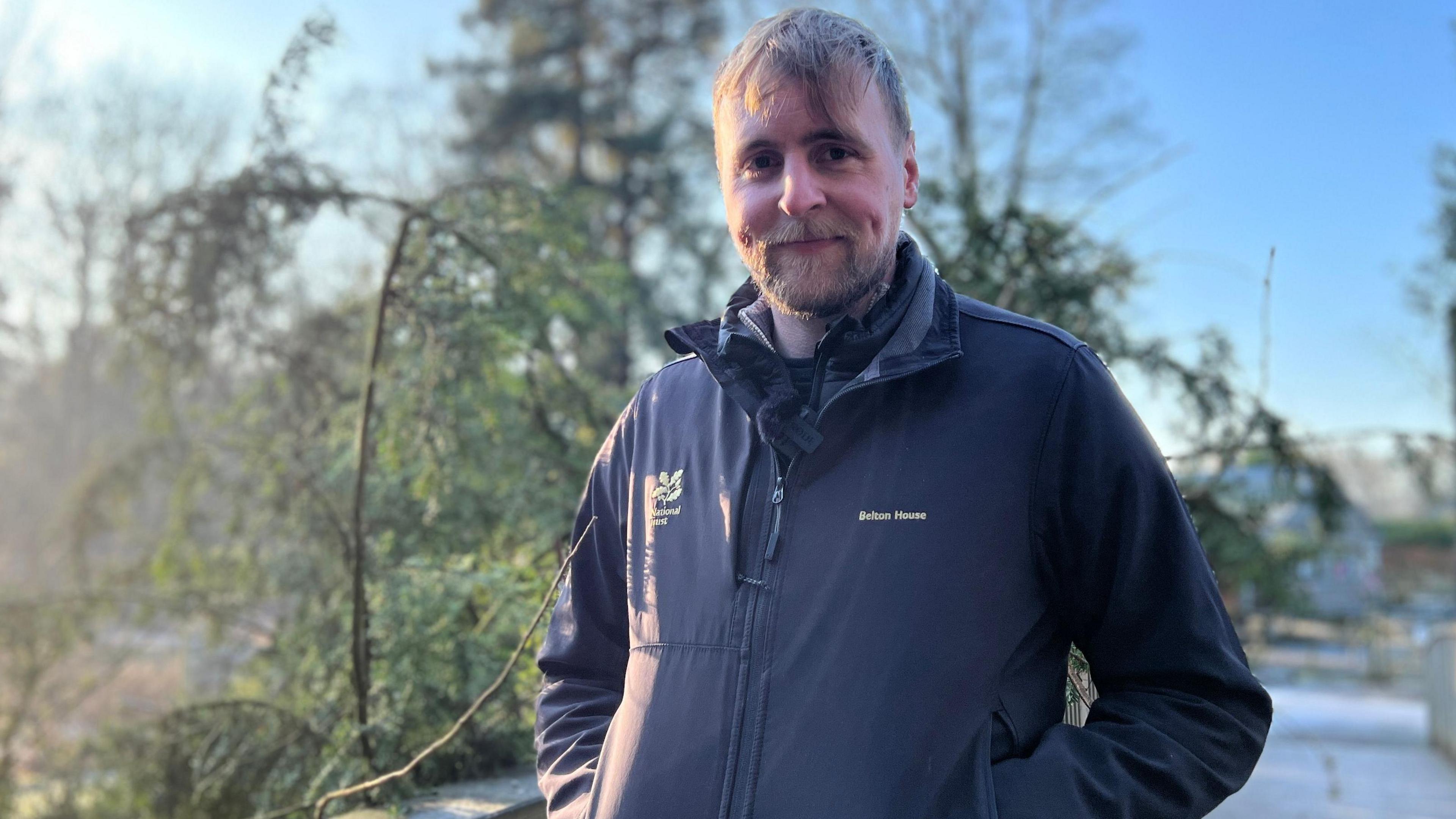 Angus Hale is wearing a black jacket and is standing in front of a tree, he is smiling.