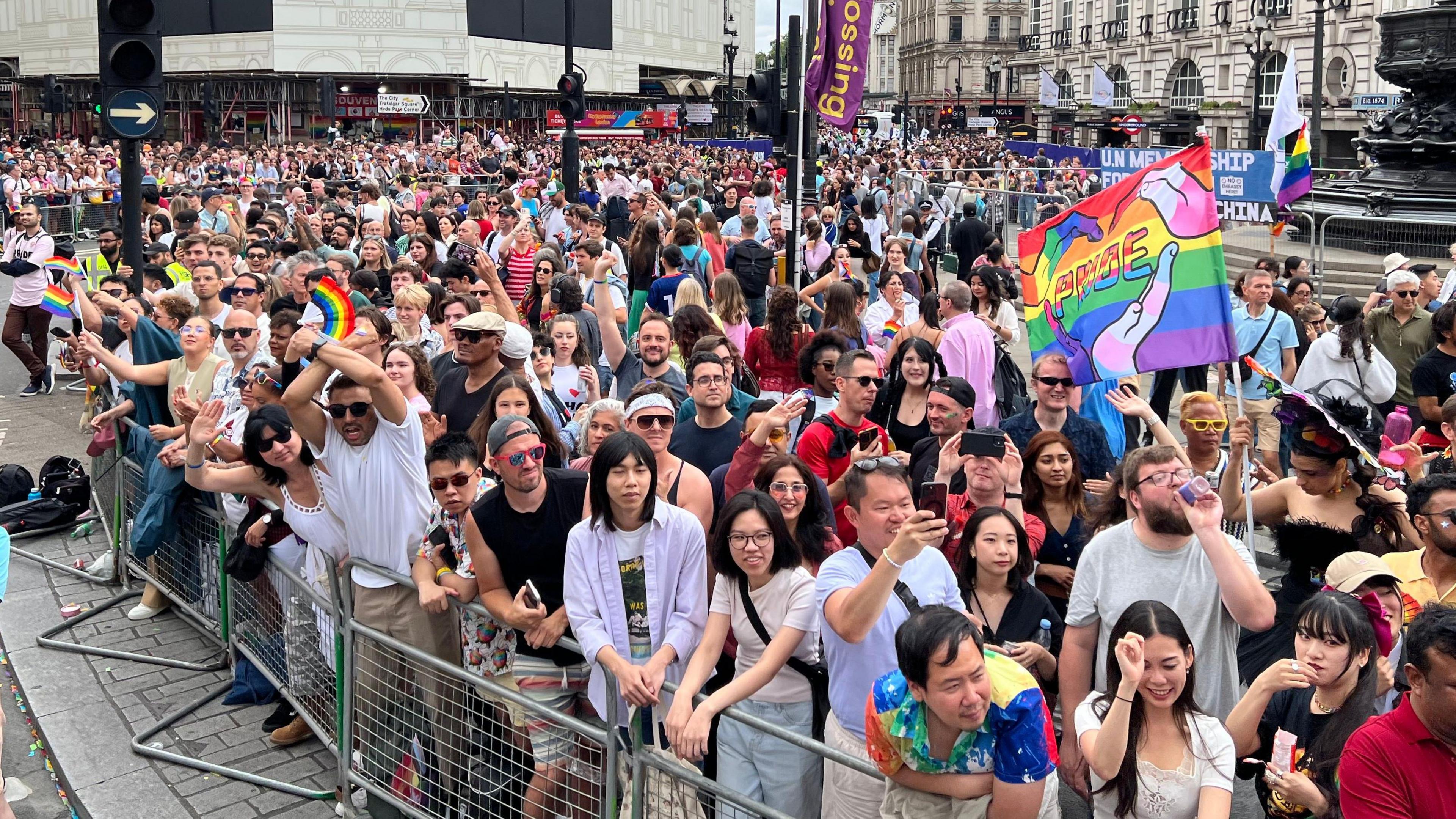 A large group of people stand behind barriers at Piccadilly Circus, some are waving LGBT+ flags while others record on their phones.