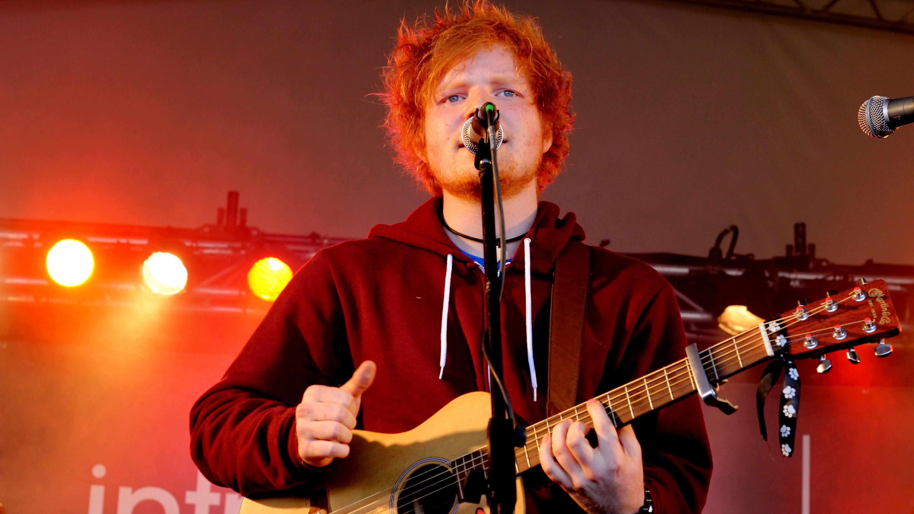 Ed Sheeran performs on the BBC Introducing Stage at Reading Festival, 2011. He has bright ginger hair and is wearing a maroon zip-up hoodie. He is singing into a microphone on a stand whilst playing an acoustic guitar.