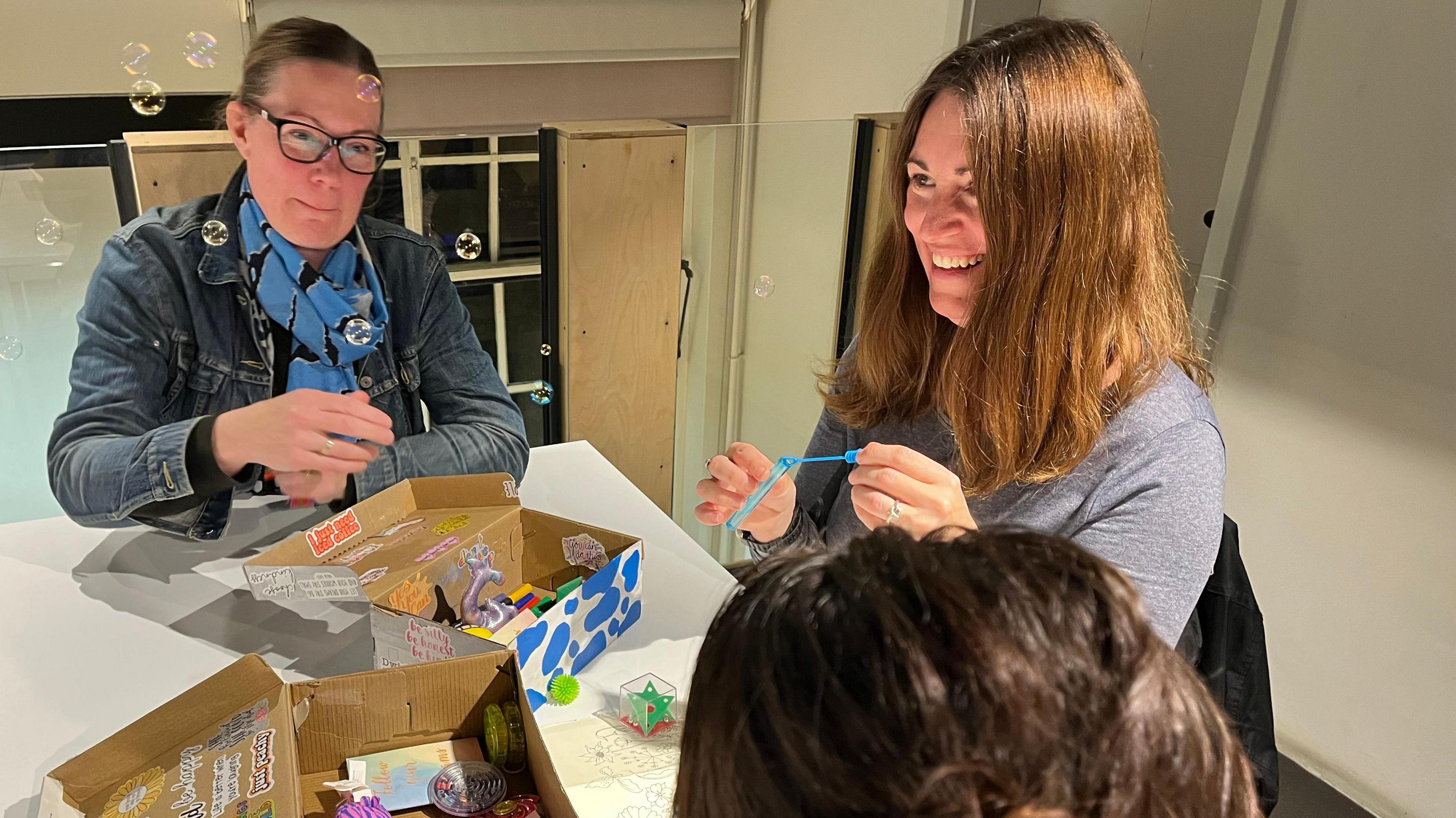 Three women sat around a table with two open shoe boxes in front of them with fidget toys inside them. The woman in the middle is blowing bubbles.  