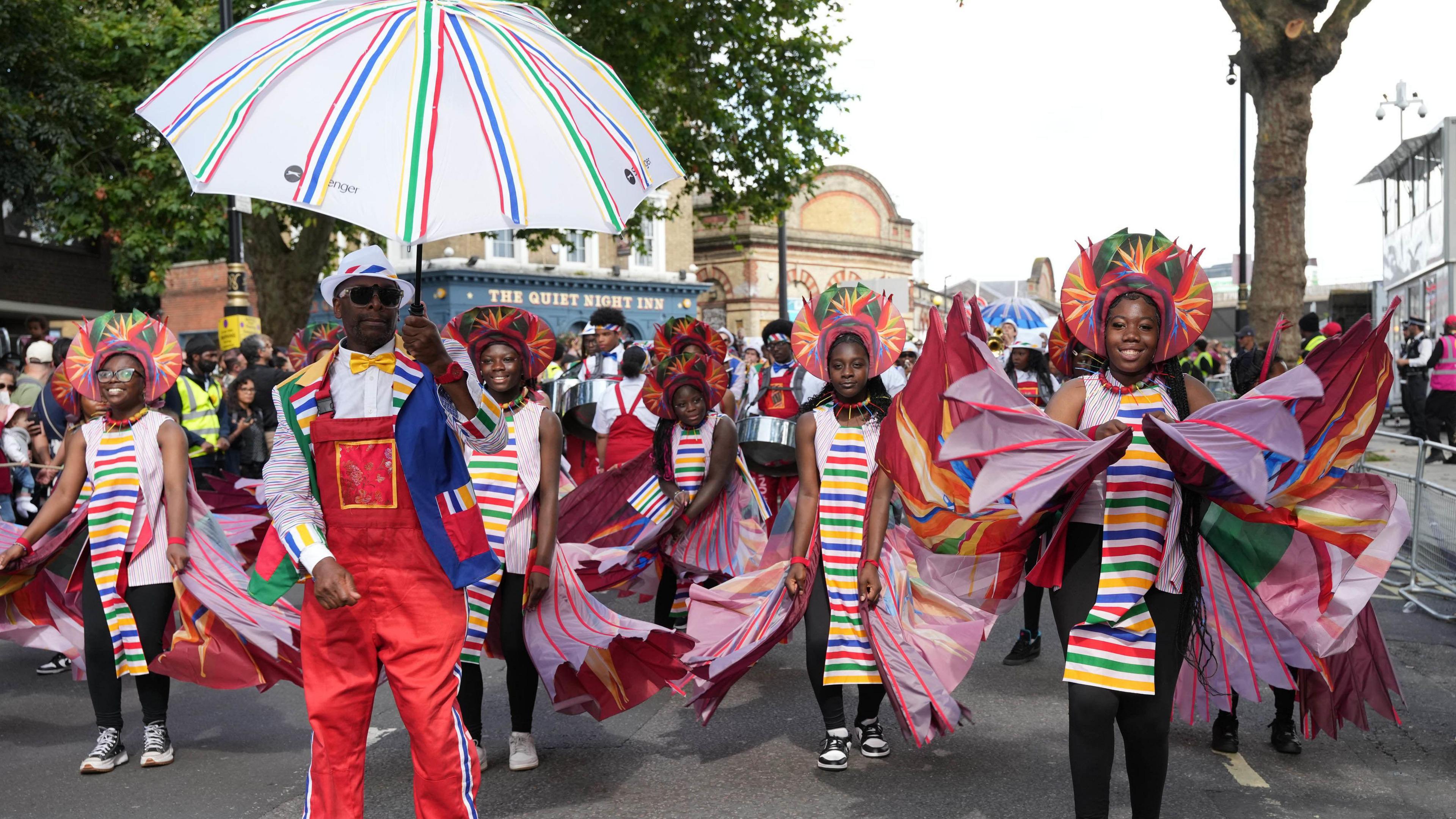 Performers in colourful, coordinated costumes dance in the street during Notting Hill Carnival, with one man holding a large striped umbrella leading a group of smiling girls wearing vibrant headpieces and wing-like fabric.