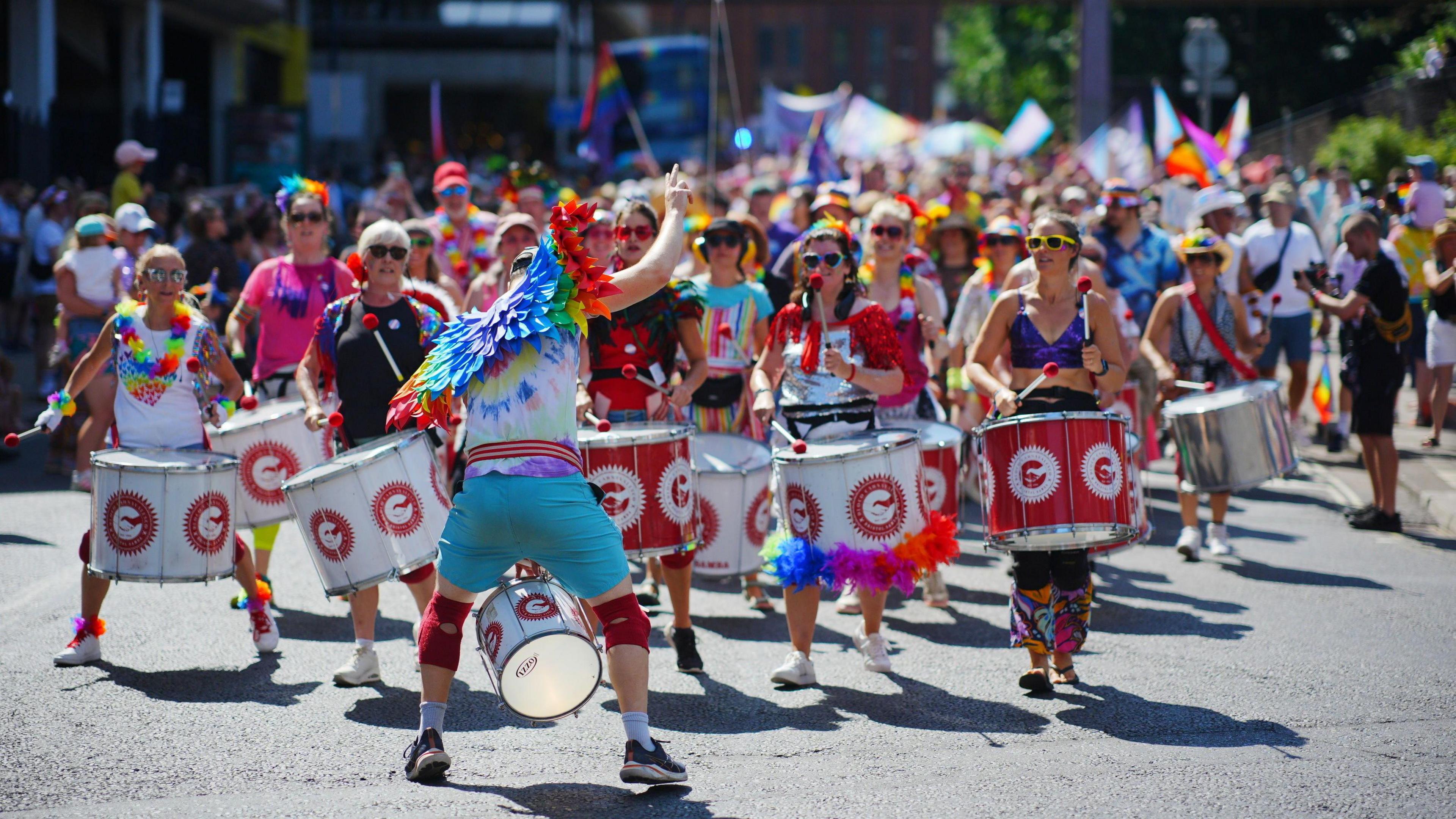 A shot looking down a large pride parade - in the foreground are a samba band, all holding red and white drums and dressed for a summer's day with rainbow accessories. The leader has his back to the camera to face and instruct the band, and wears a pair of rainbow wings