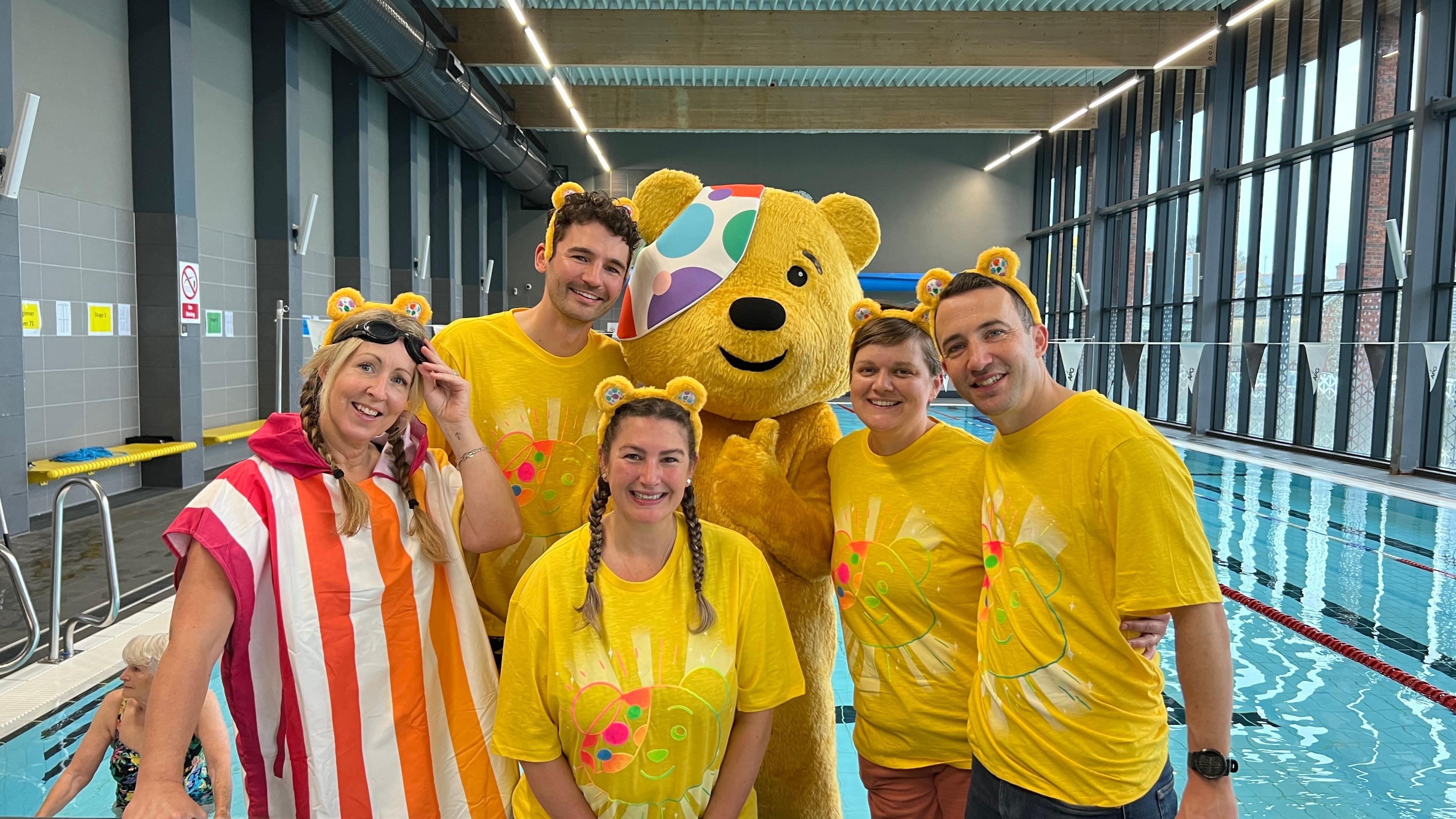 Five people and the yellow bear Pudsey looking and smiling at the camera in a swimming pool