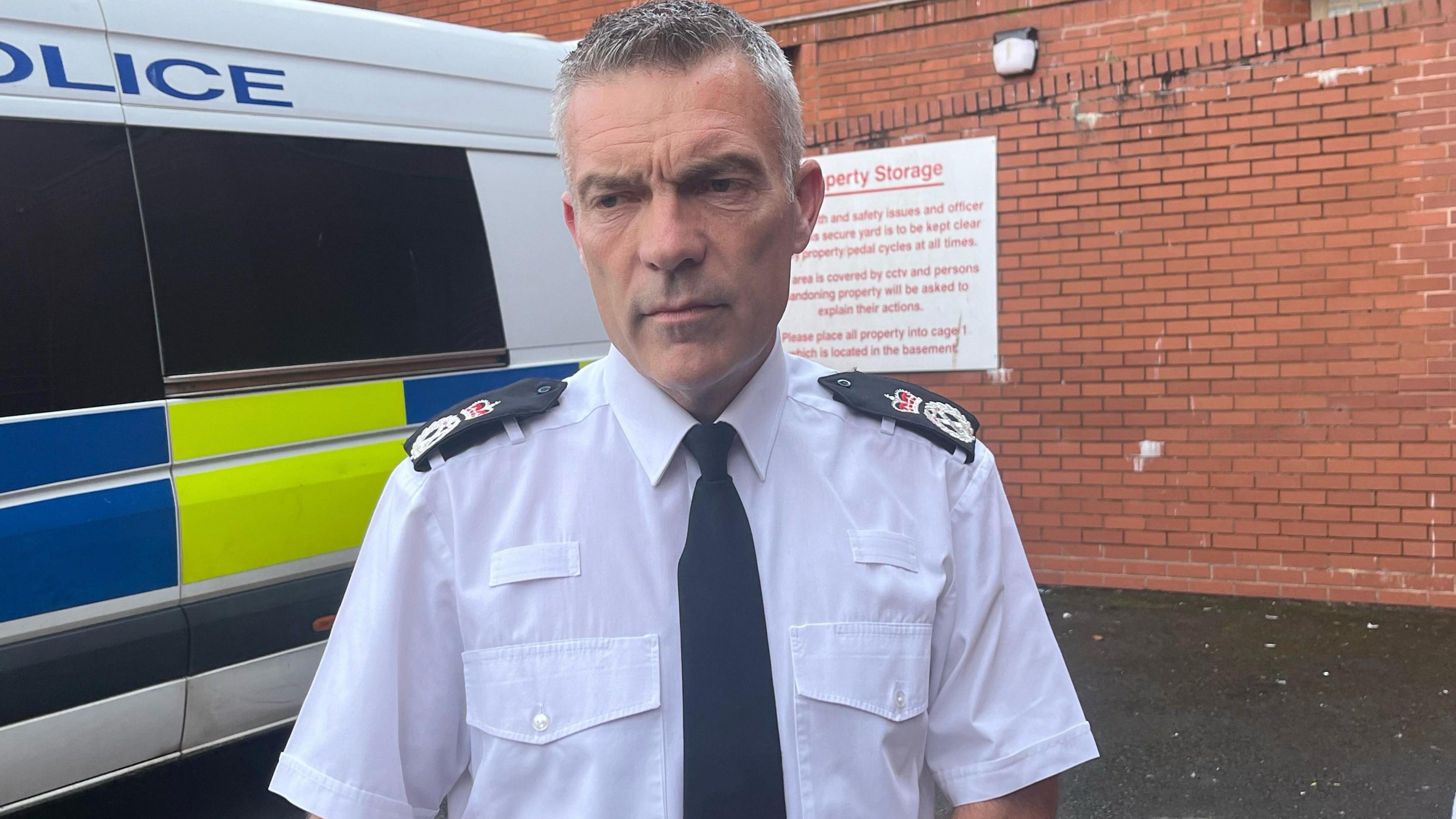 A middle-aged man with short grey hair wearing a white shirt with black epaulettes featuring a crown on the shoulders and a black tie. A police car his behind him,