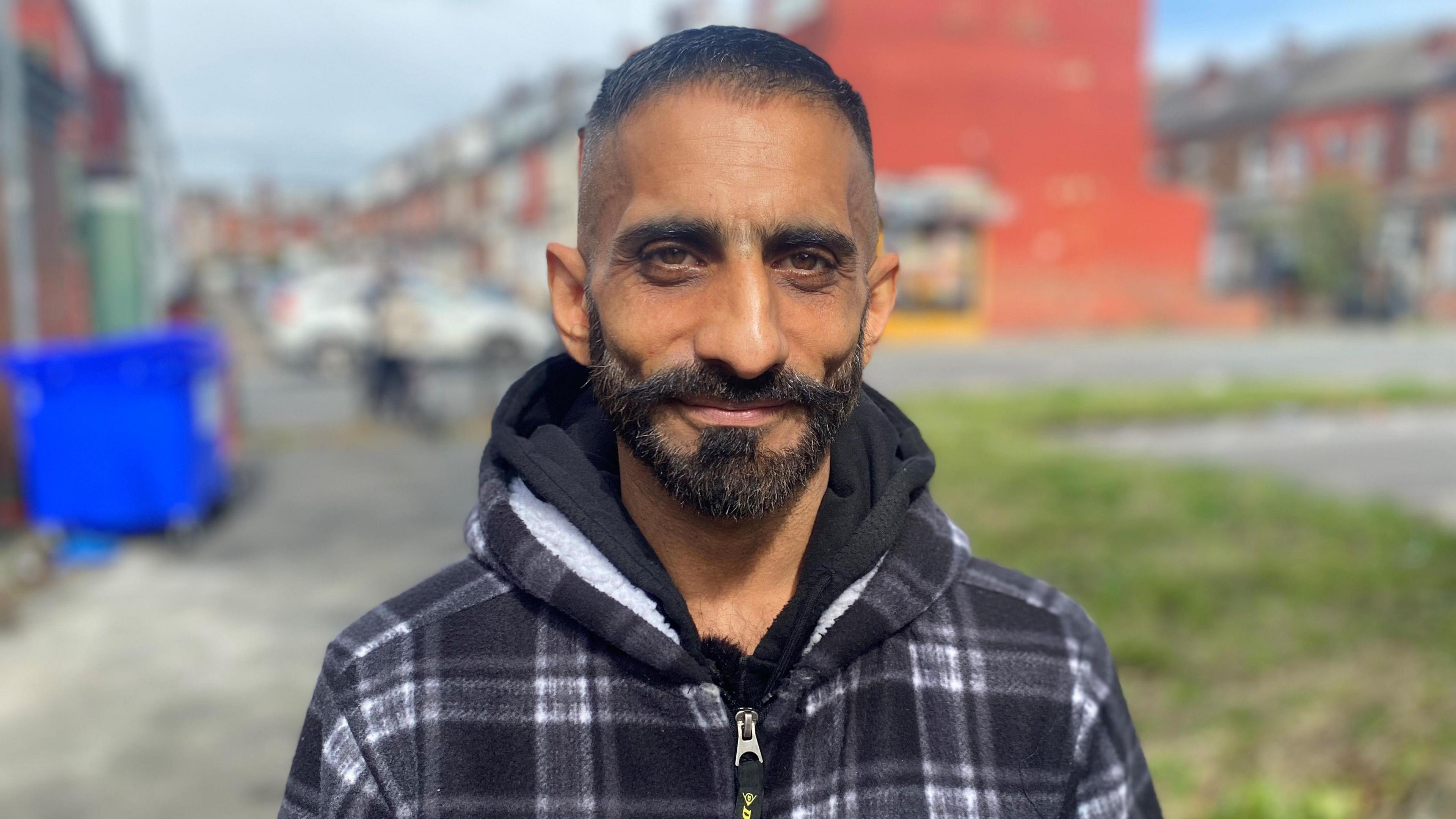 Nadsy Hussain, smiling at the camera and wearing a fleece hoodie. He has a black beard with short black hair, with Harehills residential streets in the background. 