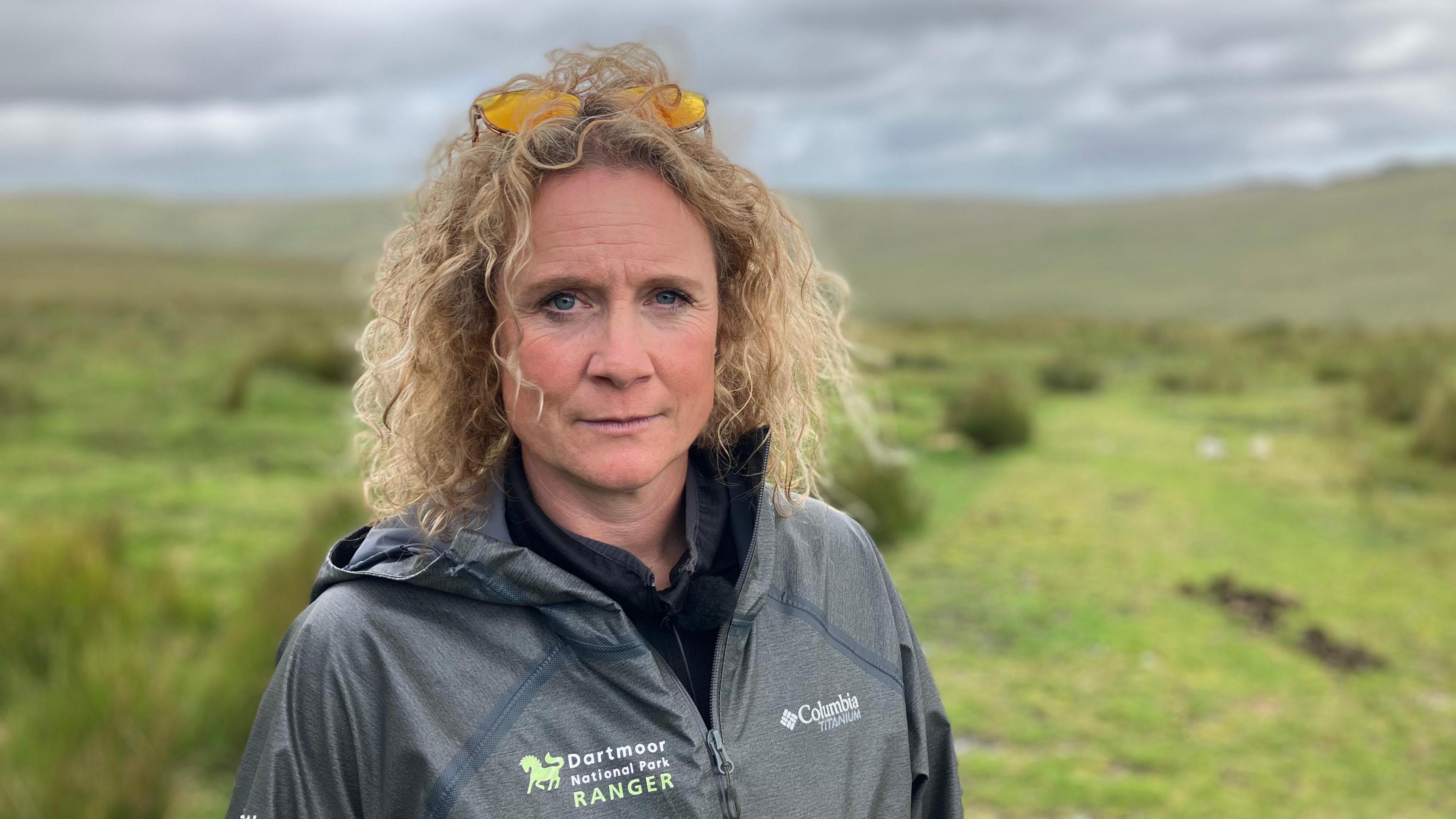 A woman with curly blond hair is standing on moorland. There are open moors, tors and livestock in the background.