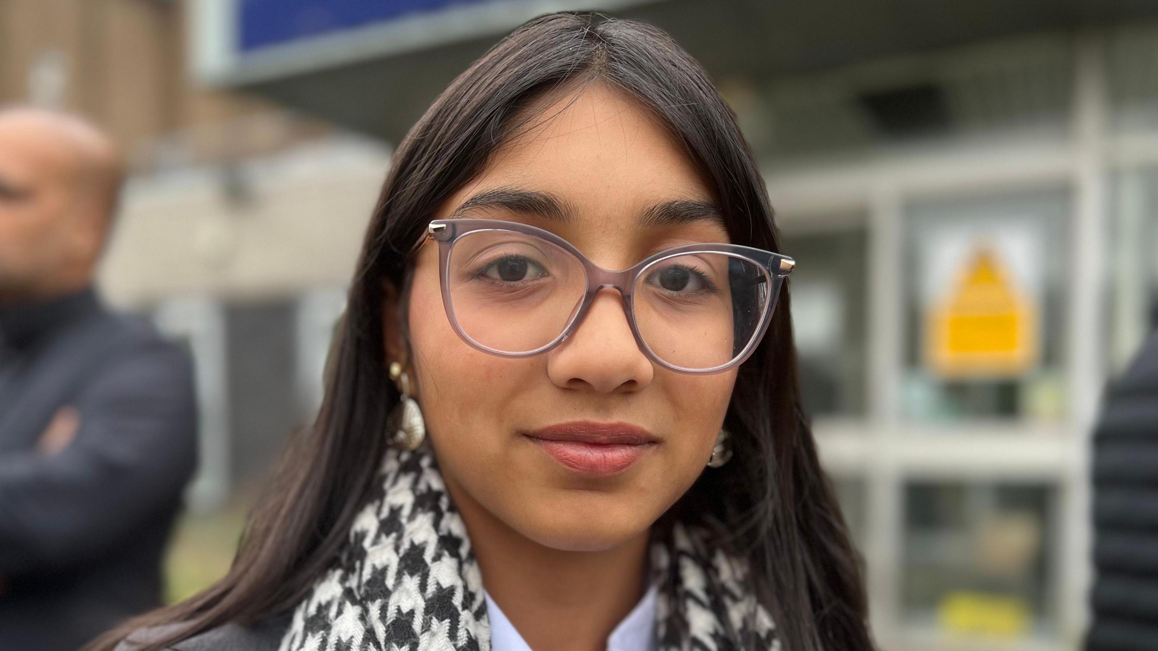 Image shows a young girls wearing glasses. 
She has a dog tooth scarf on and a shirt with a white colar. 