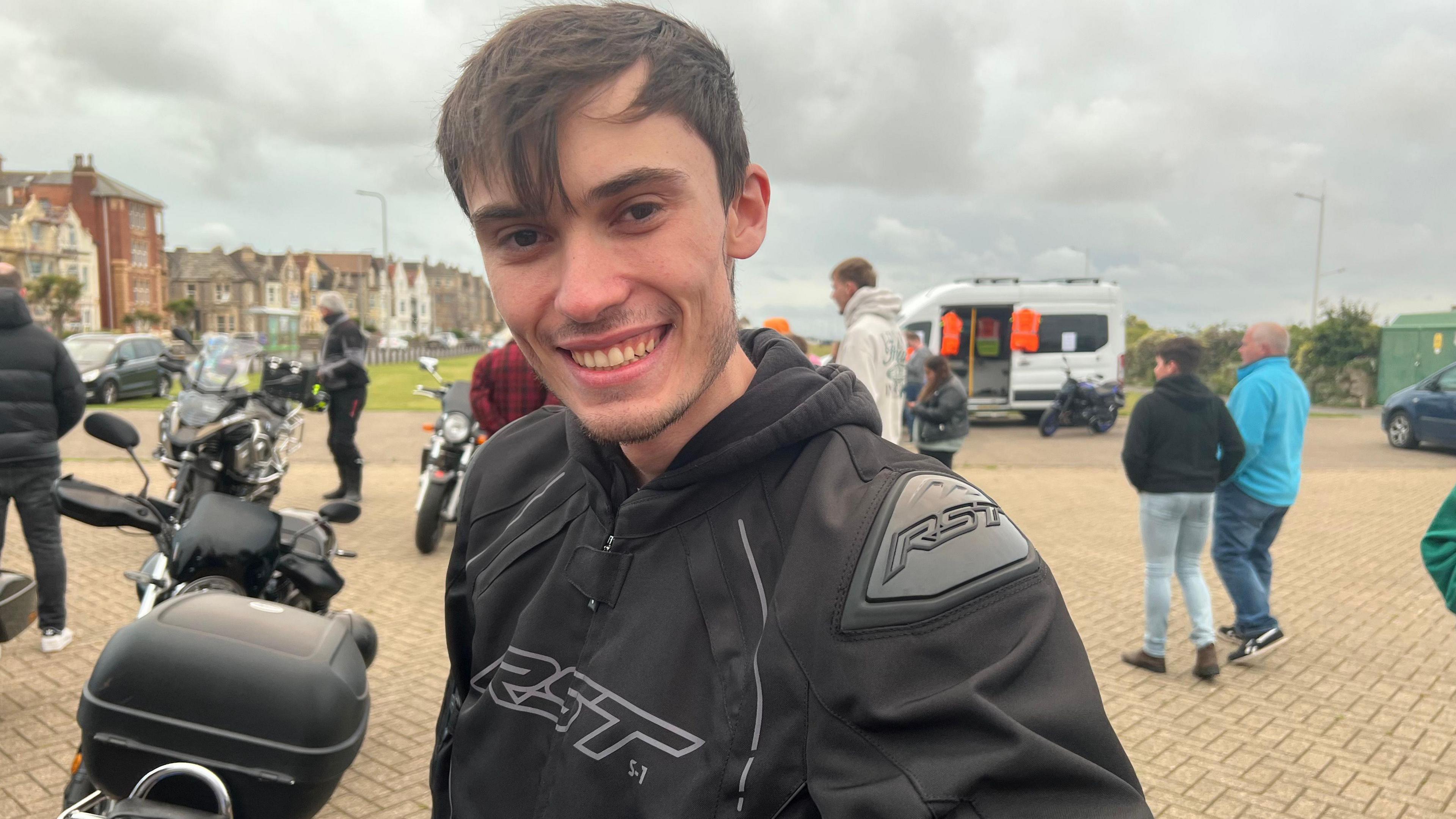 Alaistair Wearing a black hoodie with short dark hair smiling at the camera. There are people loitering in the background in a car park with a few other motorbikes in view.