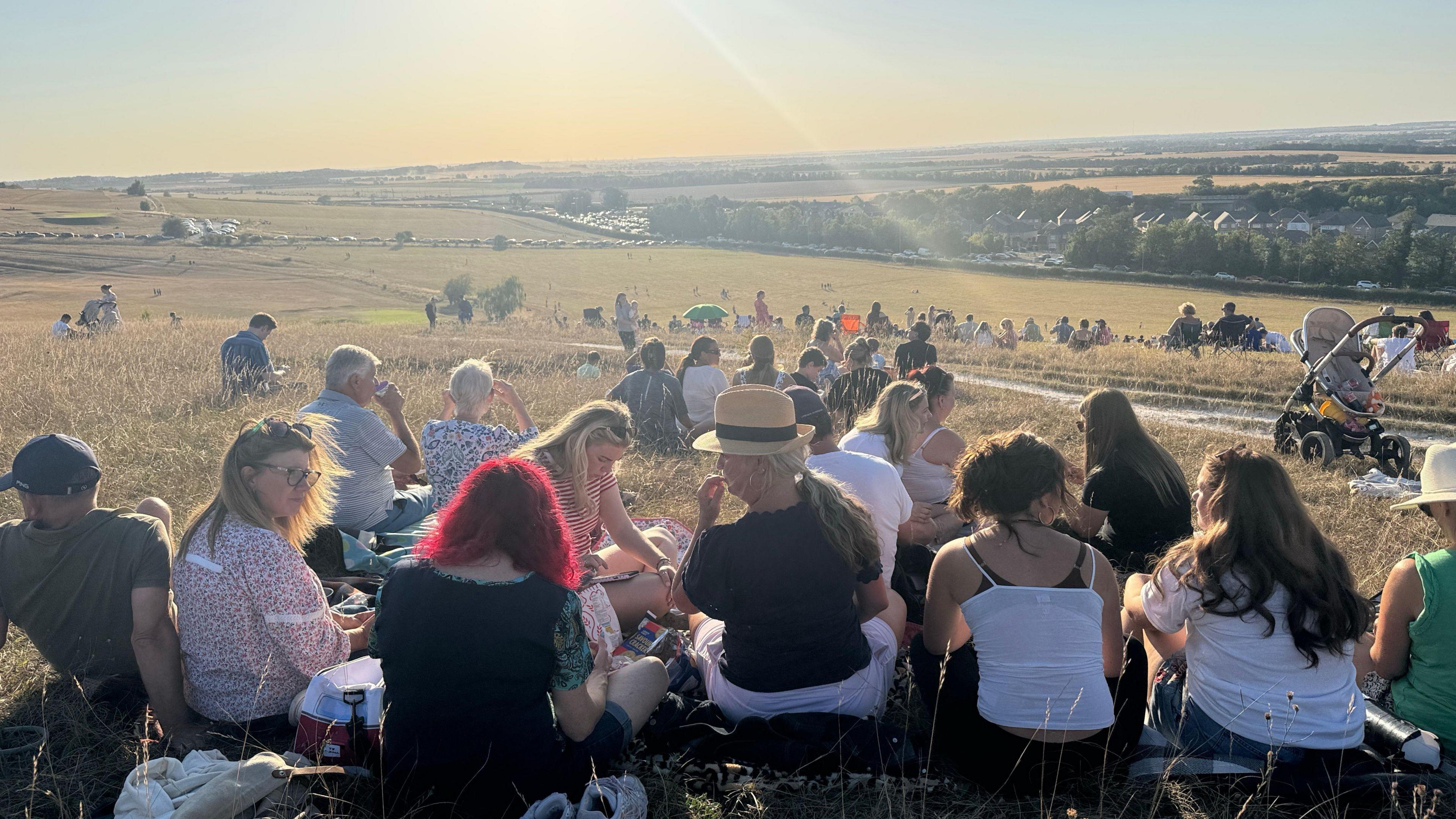 Hundreds of people sit on a sunlit hillside at Therfield Heath in Royston to watch a partial solar eclipse. The sun is bright in the sky