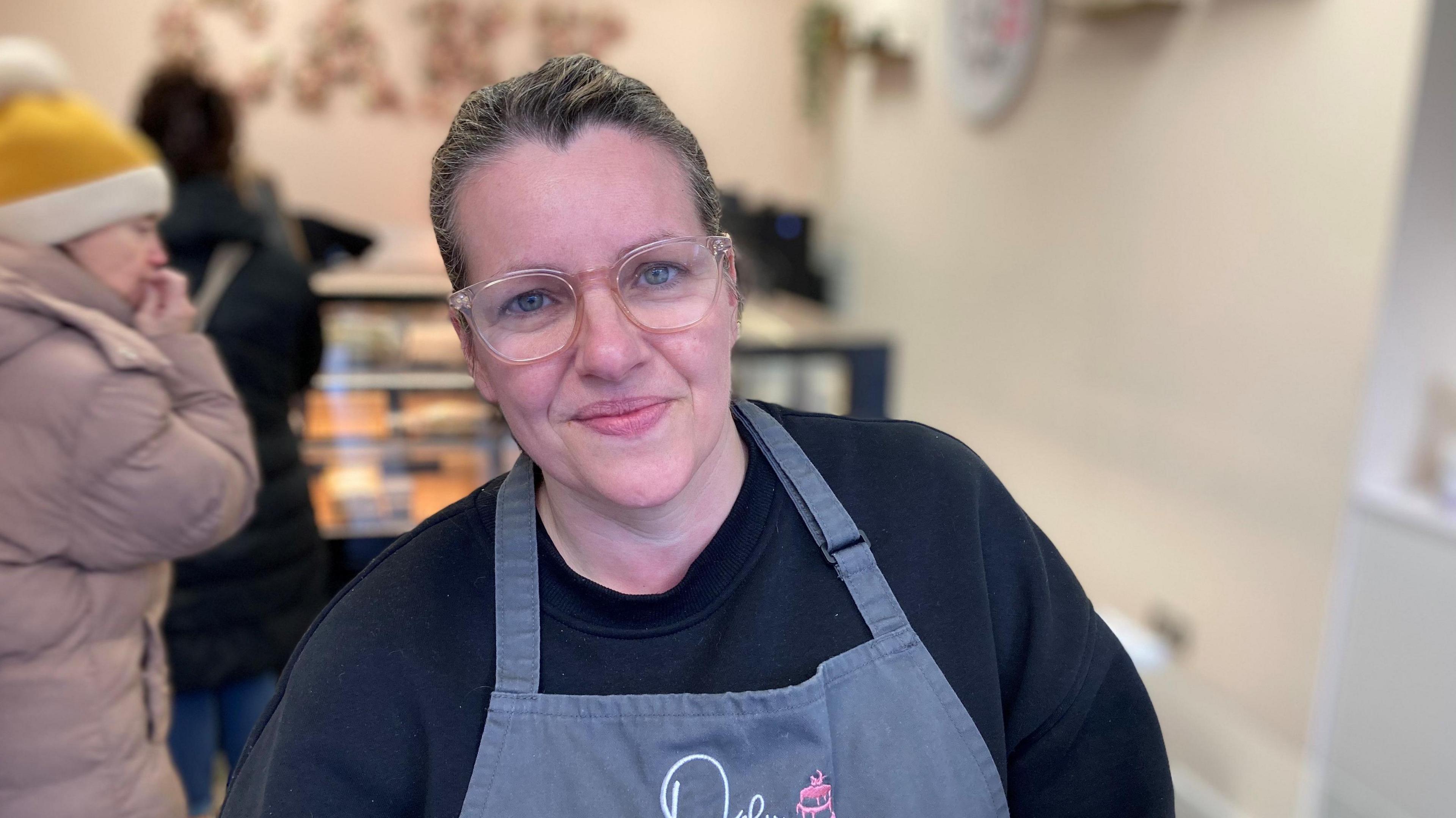 Victoria Vickery, owner of Only Crumbs sandwich and cake shop on Treorchy high street, wearing a grey apron - with customers in the background ordering food.