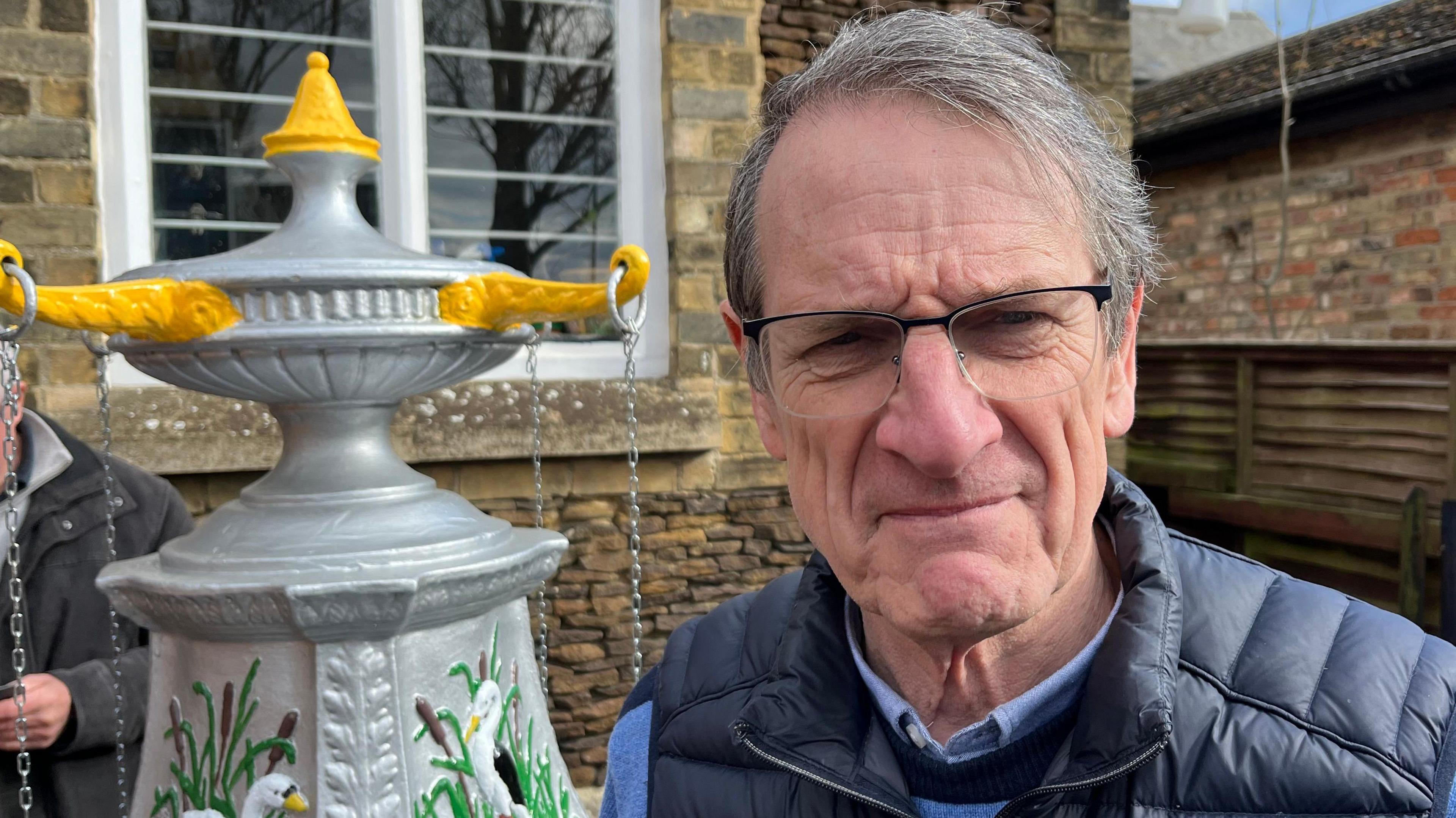 Gordon has greyish dark hair swept to the right. He is wearing a black body warmer over a blue and black striped jumper. He is wearing dark framed glasses and the water feature is on the left behind him.