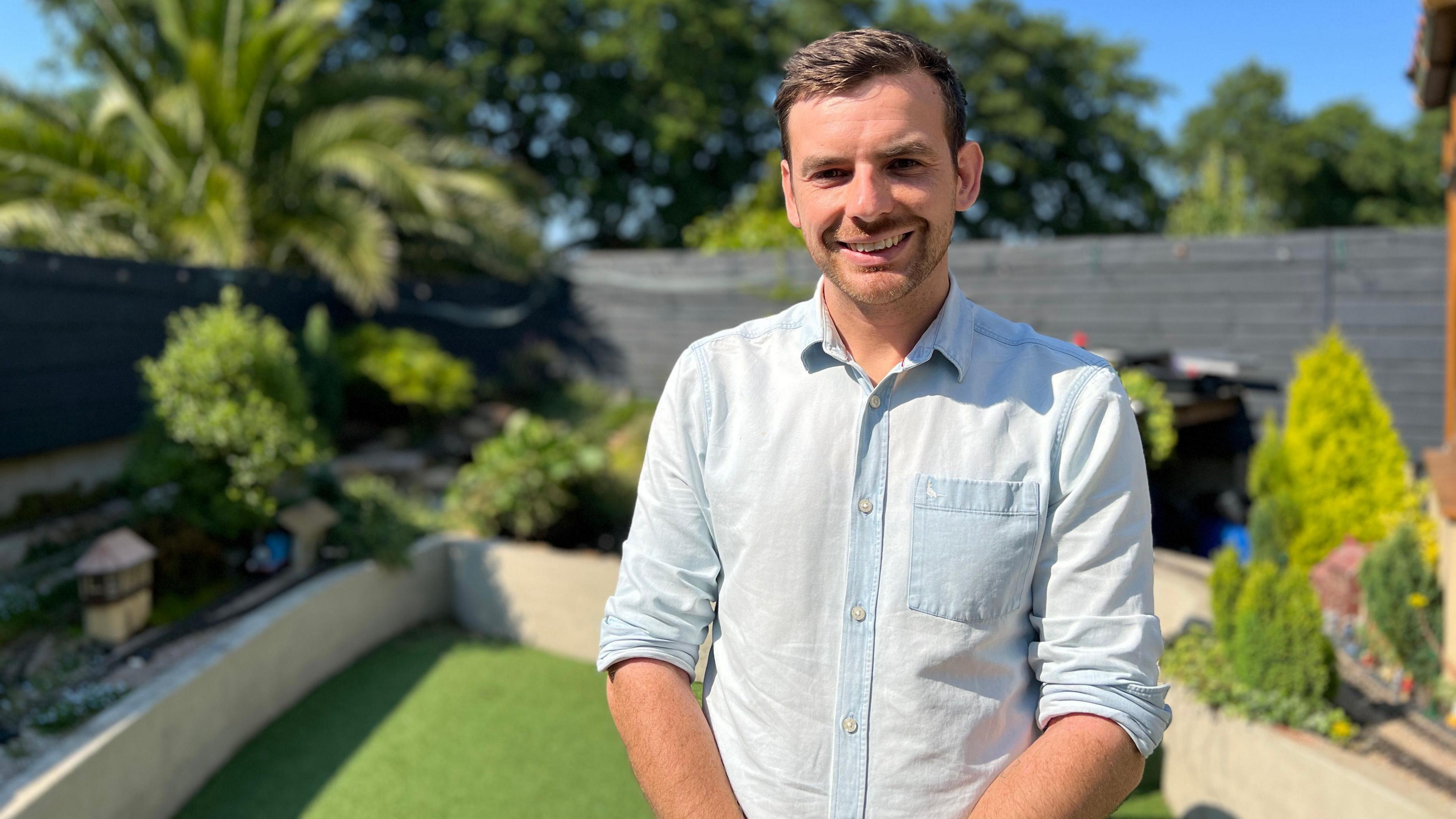 Curtis stands in the middle of the garden smiling at the camera with model trains behind him