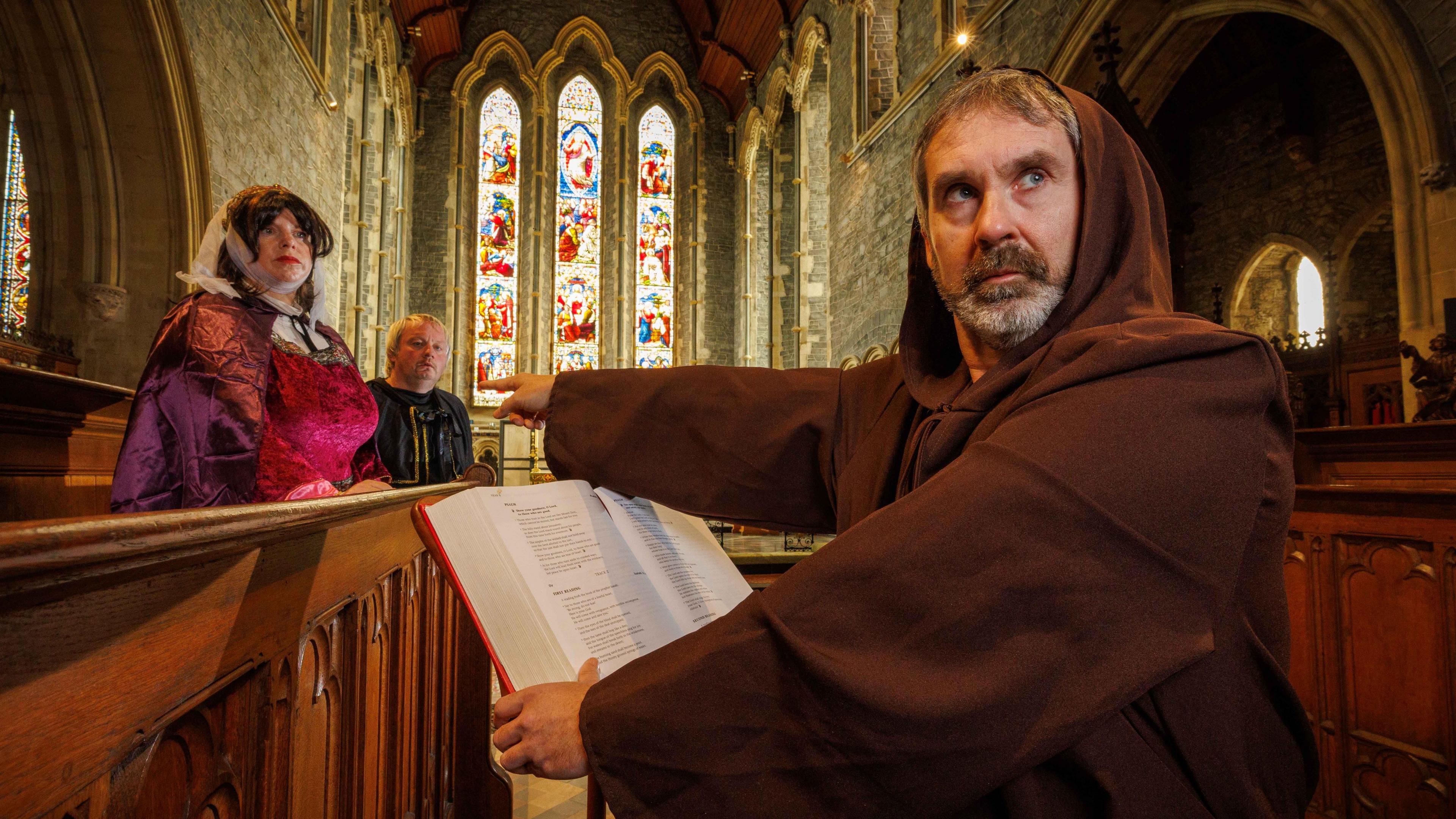 a man in a monk's cowl, holding a red book, pointing at a woman who is wearing medieval dress. She looks alarmed. They are both inside a church and its stained glass windows are visible in the background