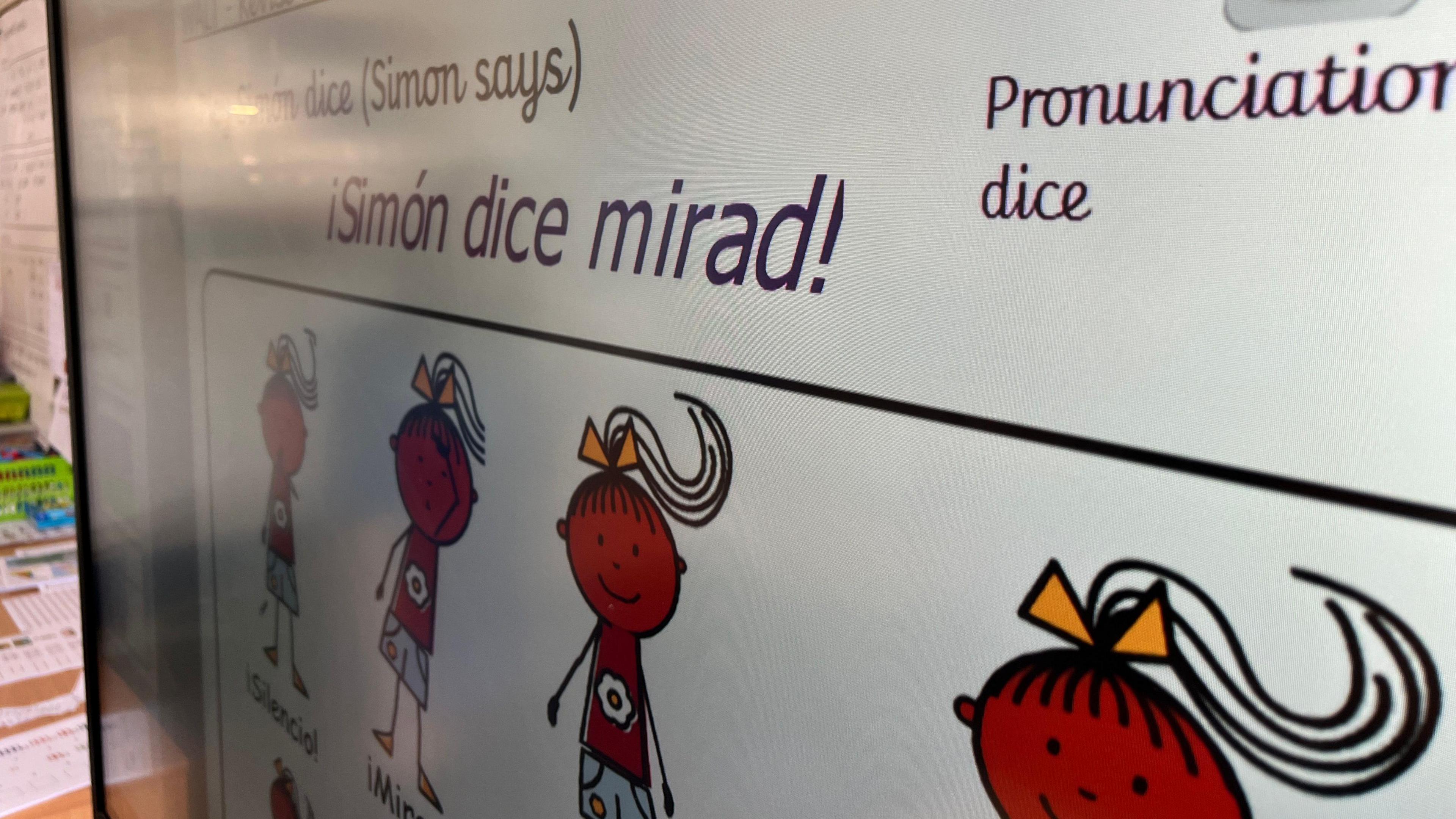 A close up picture of an electronic whiteboard in school. It shows hand drawn figures of a girl with a bow in her hair. she is pointing to different areas of her body and giving Simon Says instructions in Spanish.