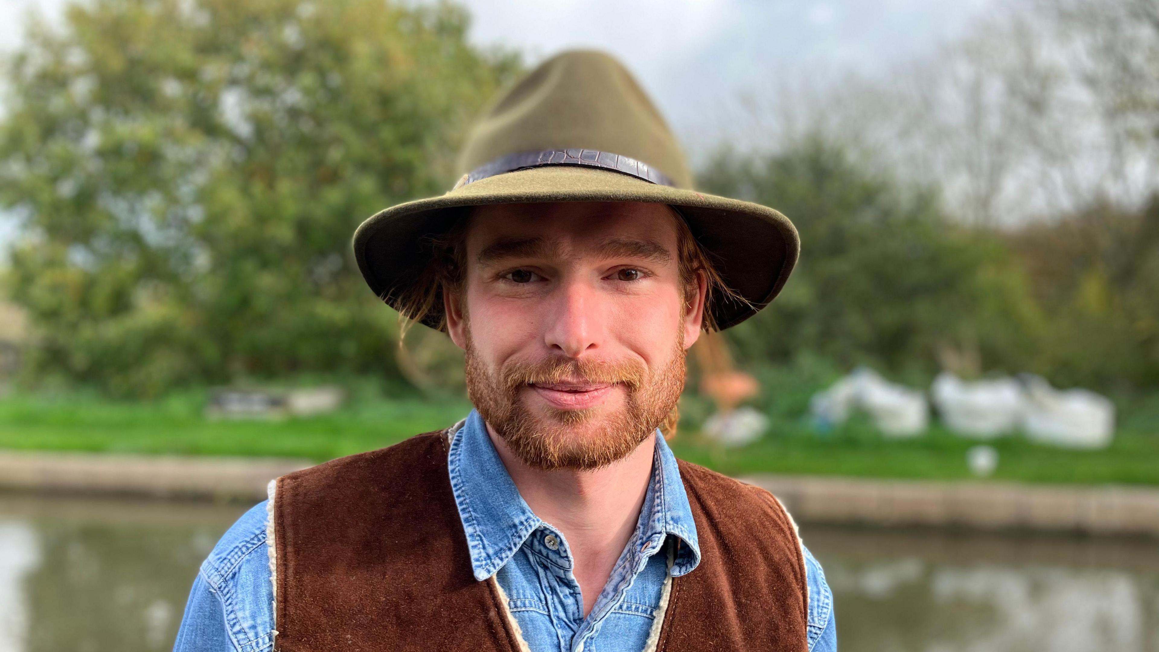 Xavier is stood facing the camera with a slight smile, wearing a green fedora and denim shirt with a brown waistcoat over the top. He is stood in front of the canal.