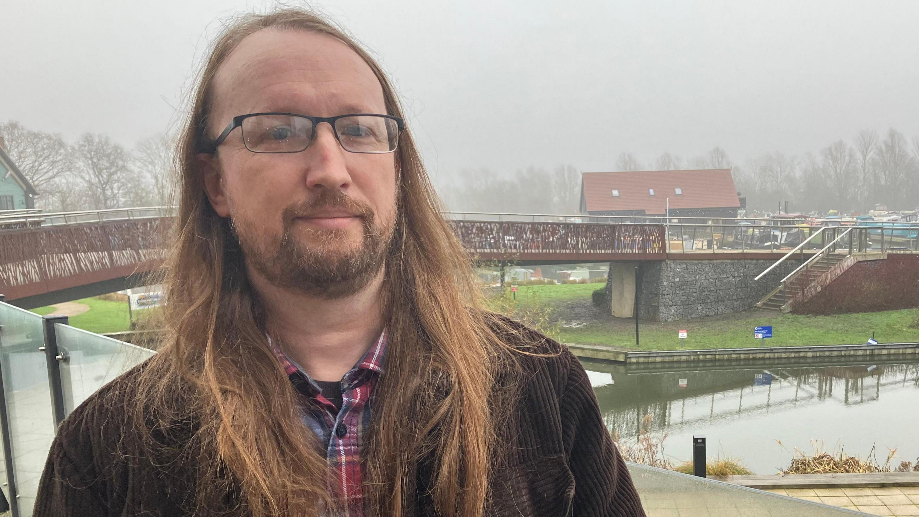 A picture of a man with long brown hair that goes down to this stomach, he also has glasses and a beard and is wearing a black jacket with a lumberjack shirt.