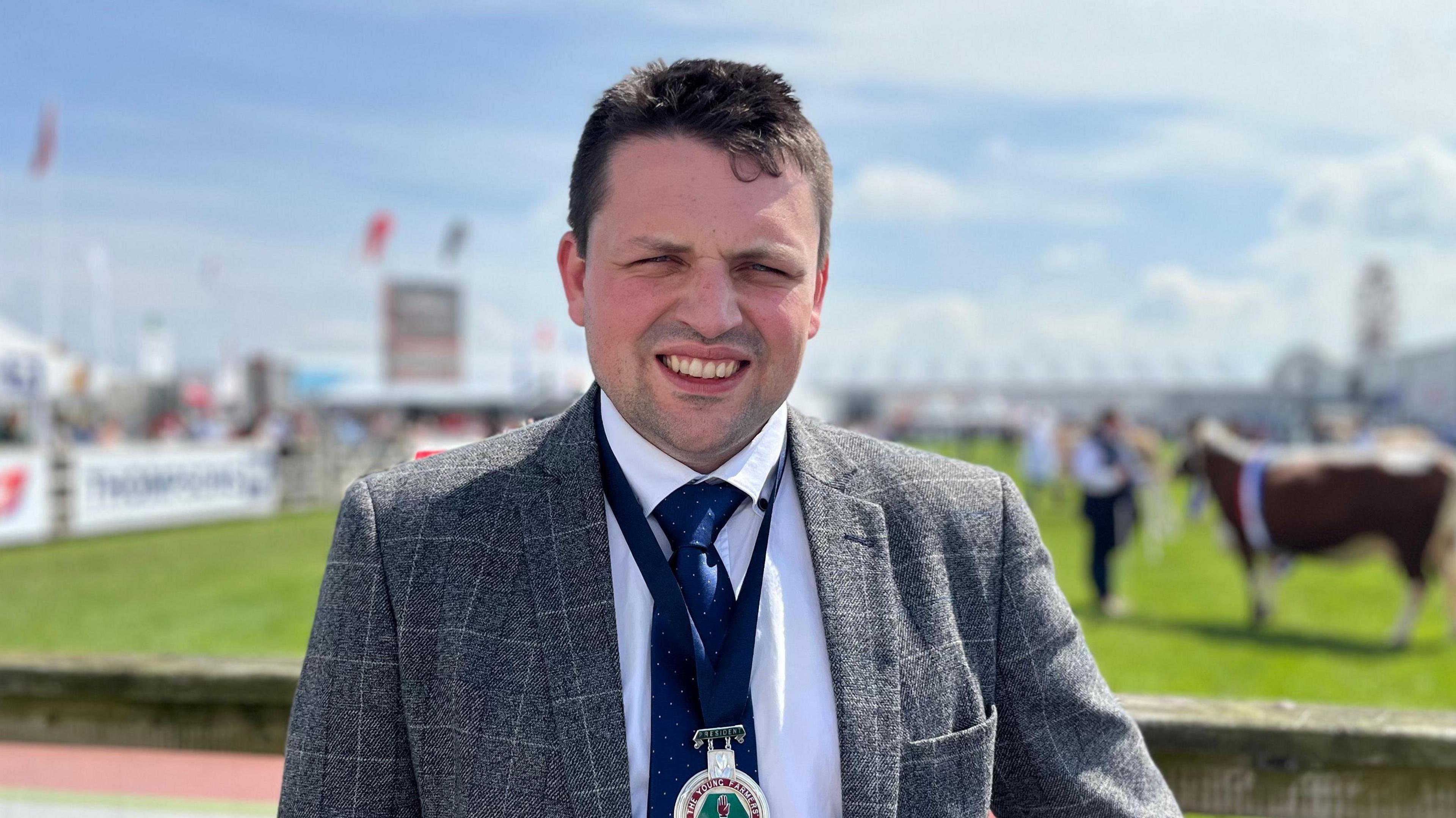 A man smiling into the camera. He has dark hair and is wearing a grey suit, with a blue tie and a blue medal around his neck. He is at a agriculture competition, there is a prize bull in the background. 