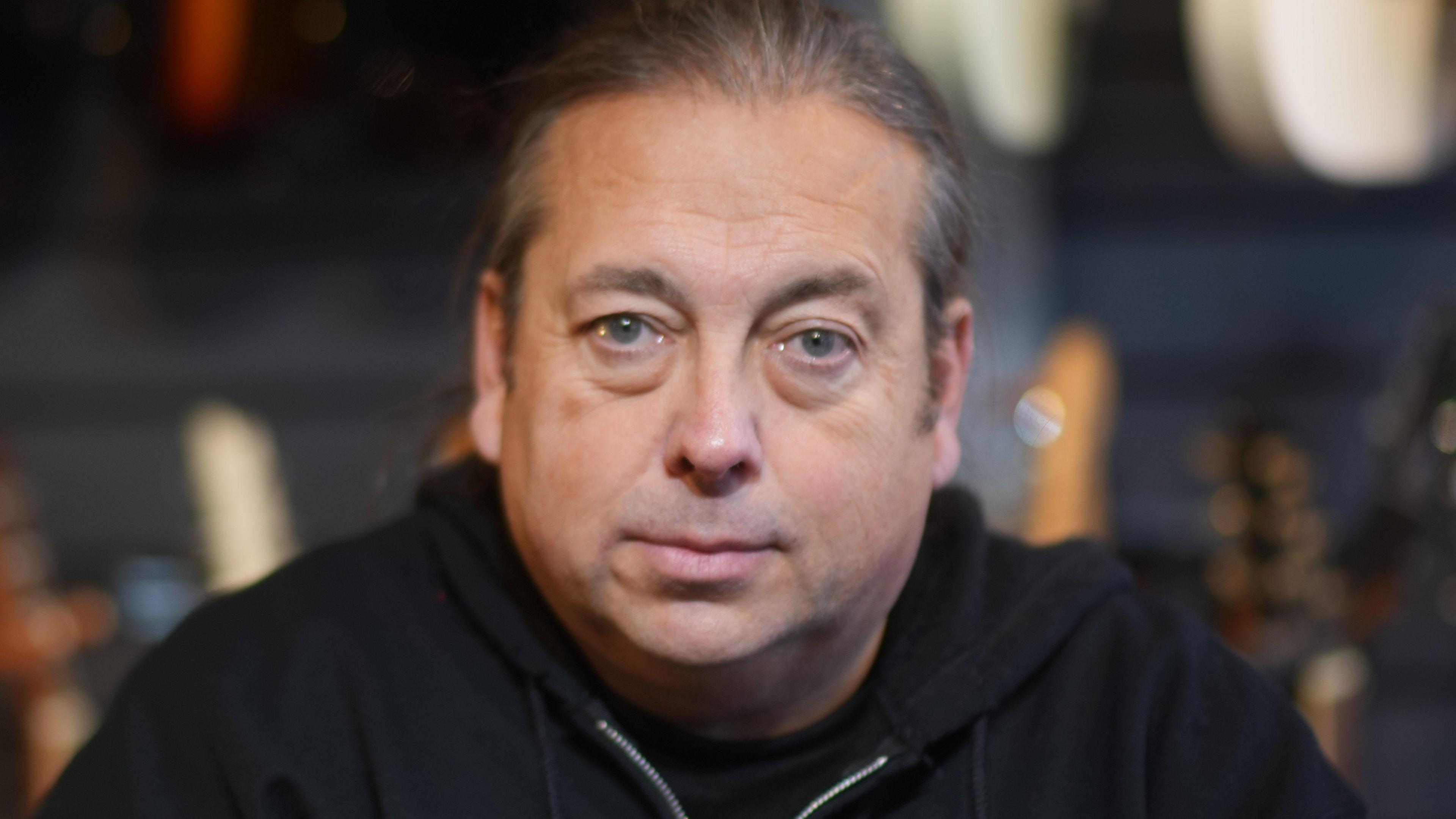 A man with long grey hair that is tied back smiles at the camera. He is wearing a black hoodie with a black t-shirt underneath. 