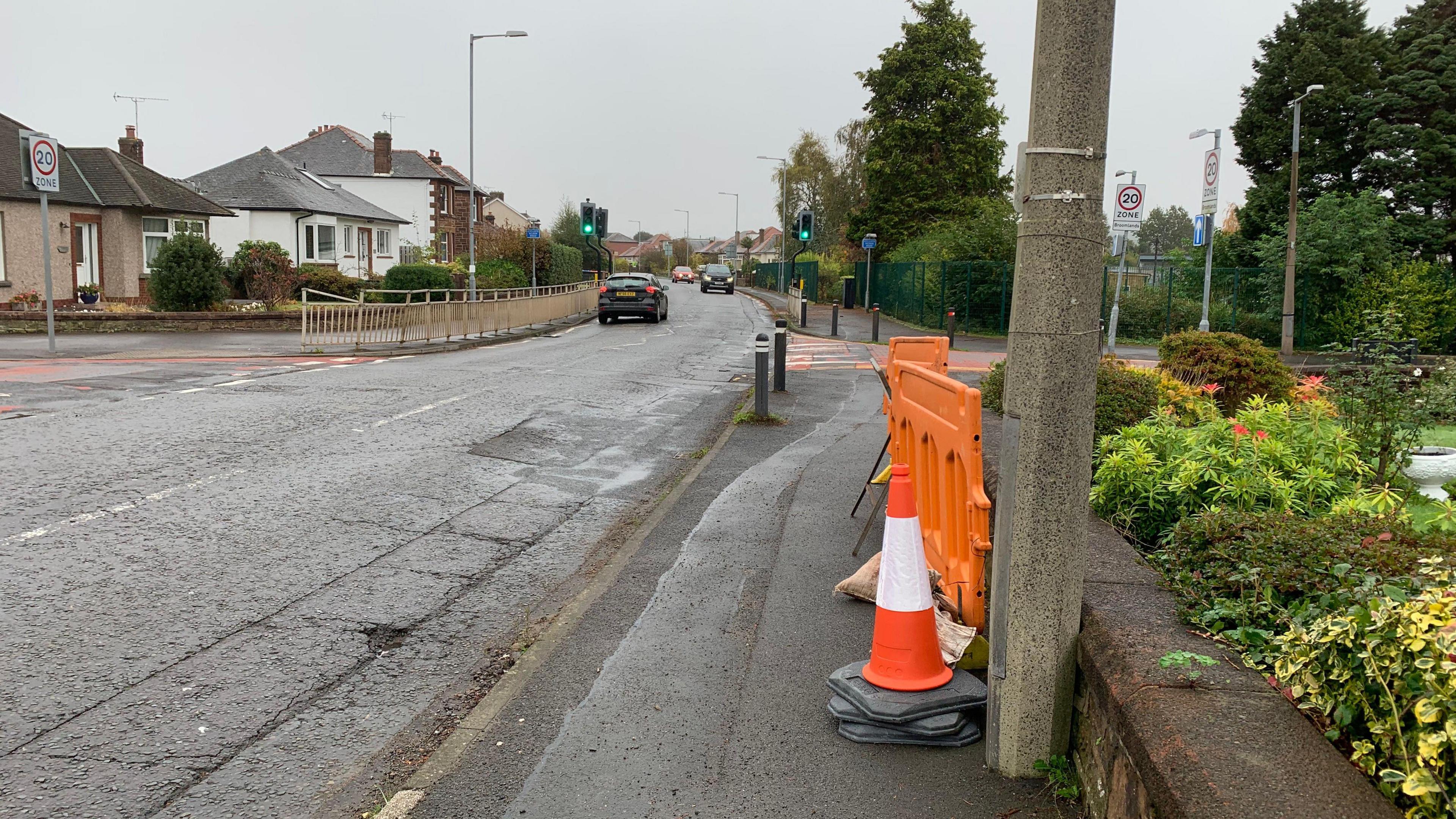 A traffic cone and barriers tidied away on a pavement. There are a couple of cars on the residential street on a very grey day. 