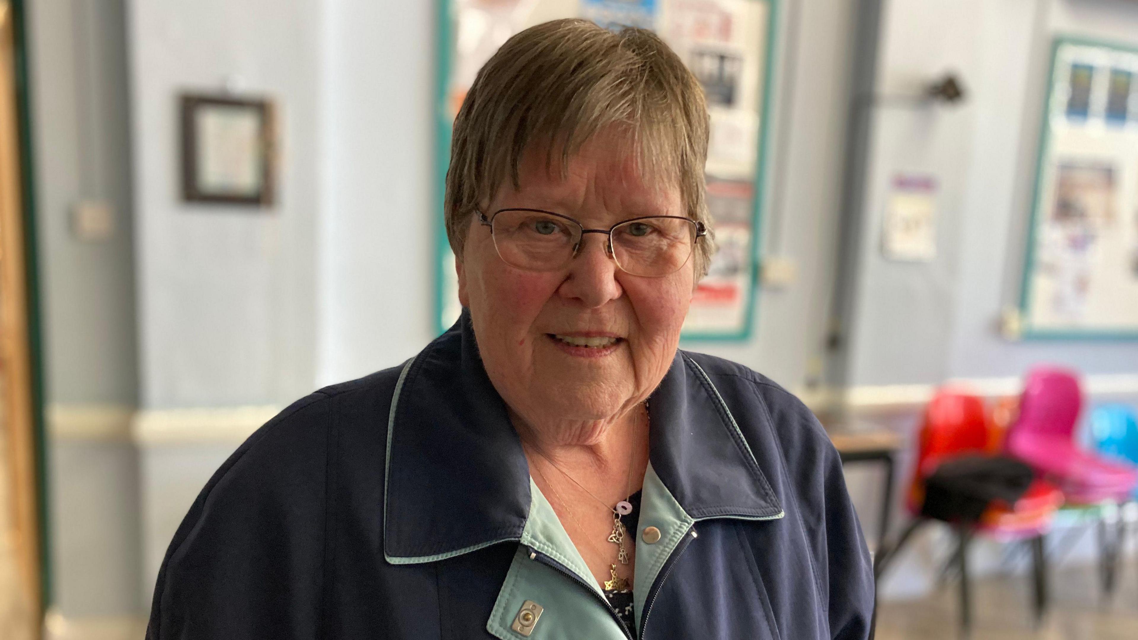 Head and shoulders image of Wendy standing in a community meeting room. She has short brown hair, glasses and is wearing a navy overcoat.