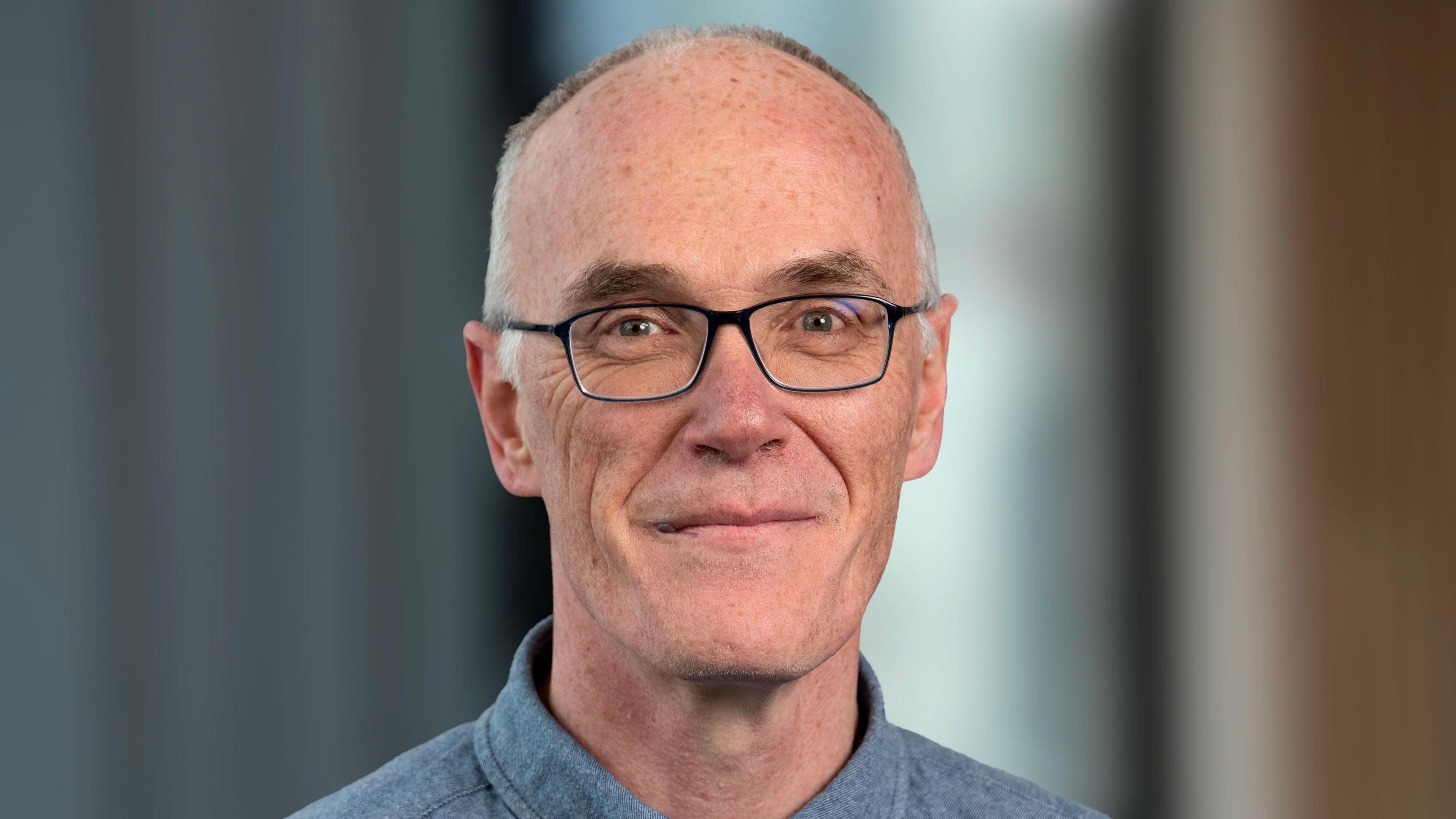 Dr Eoin Magennis is smiling at the camera in a lecture hall wearing a pair of dark framed glasses. He has short grey/white hair and is wearing a blue/grey shirt.